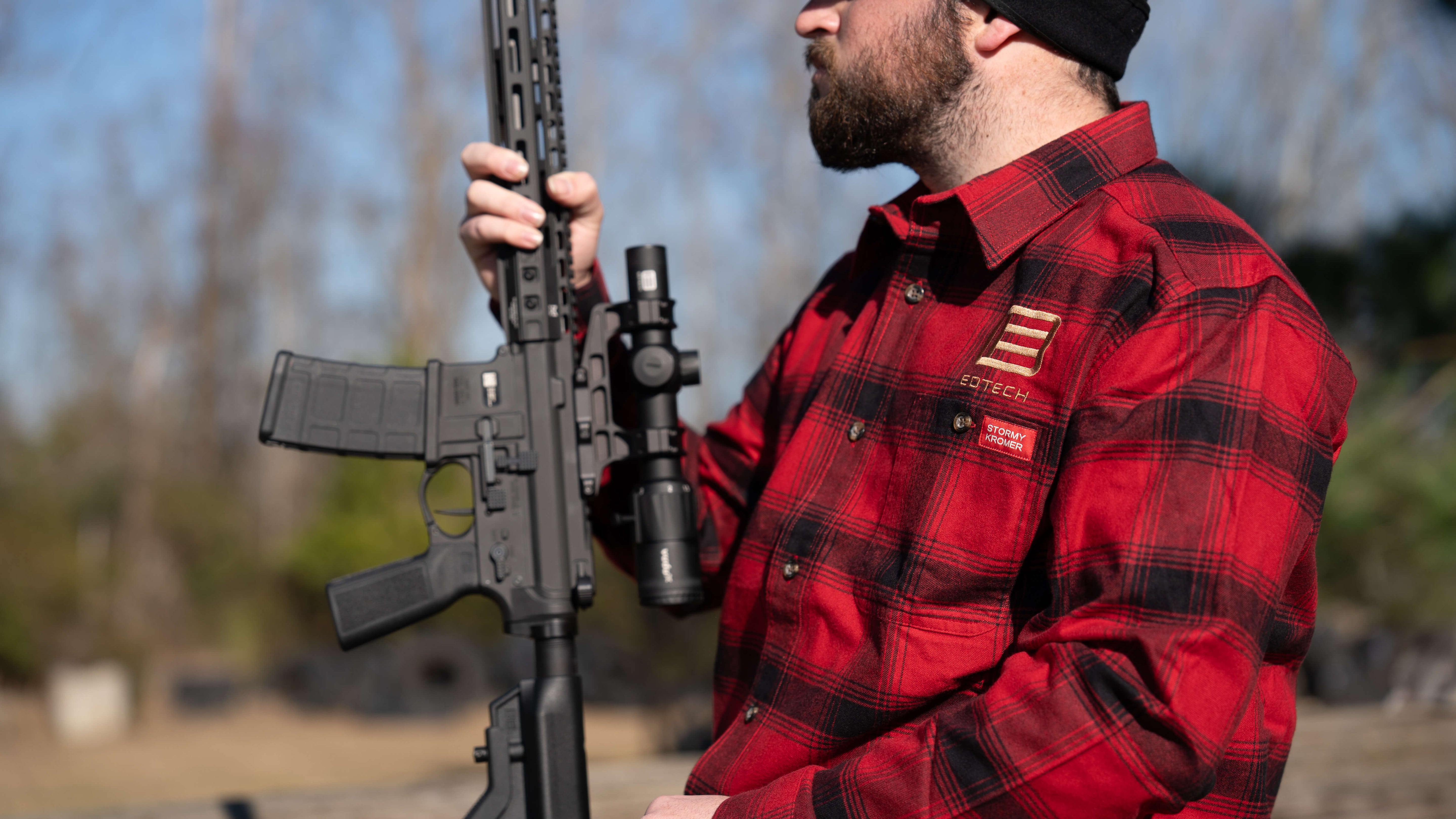 Person wearing a red plaid shirt with EOTECH logo holding an rifle with Vudu optic outdoors
