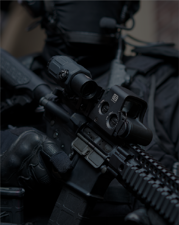 Close-up of a rifle with black EOTECH HWS optic and magnifier and tactical gear, likely in a military or law enforcement context.