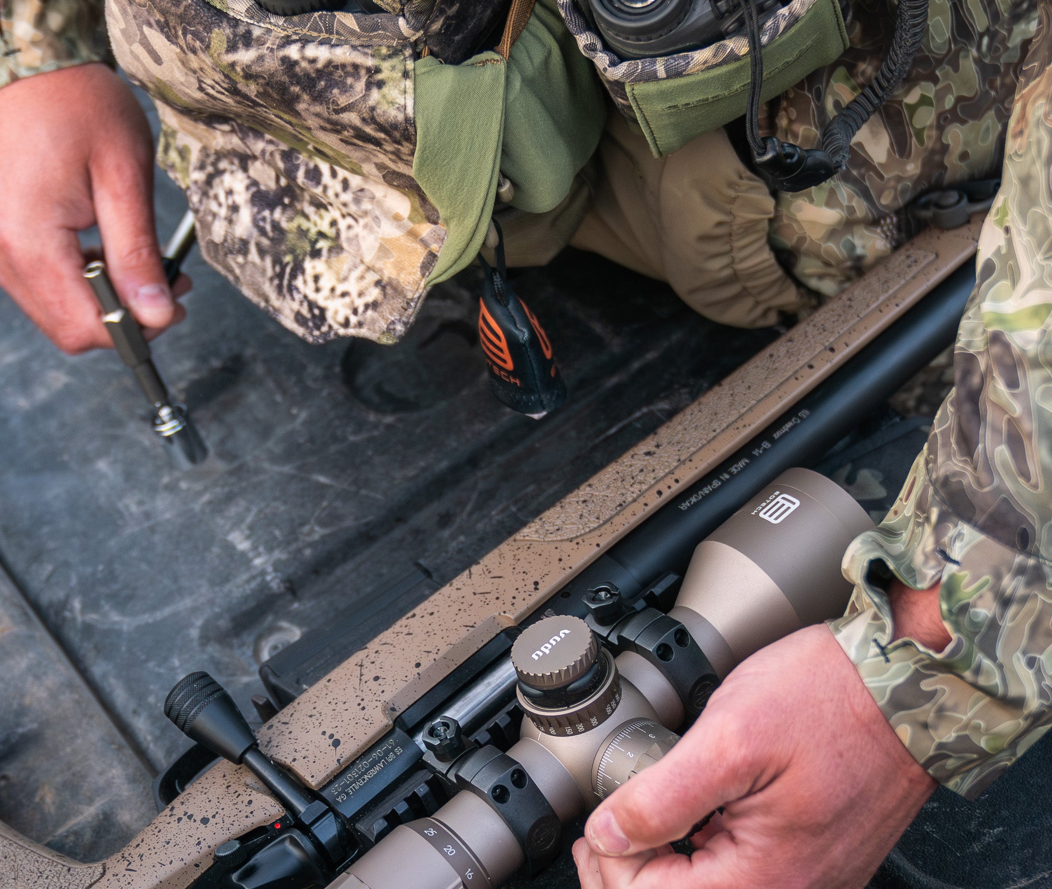 Person in camouflage gear adjusting a rifle with a EOTECH Vudu tan scope in an outdoor setting