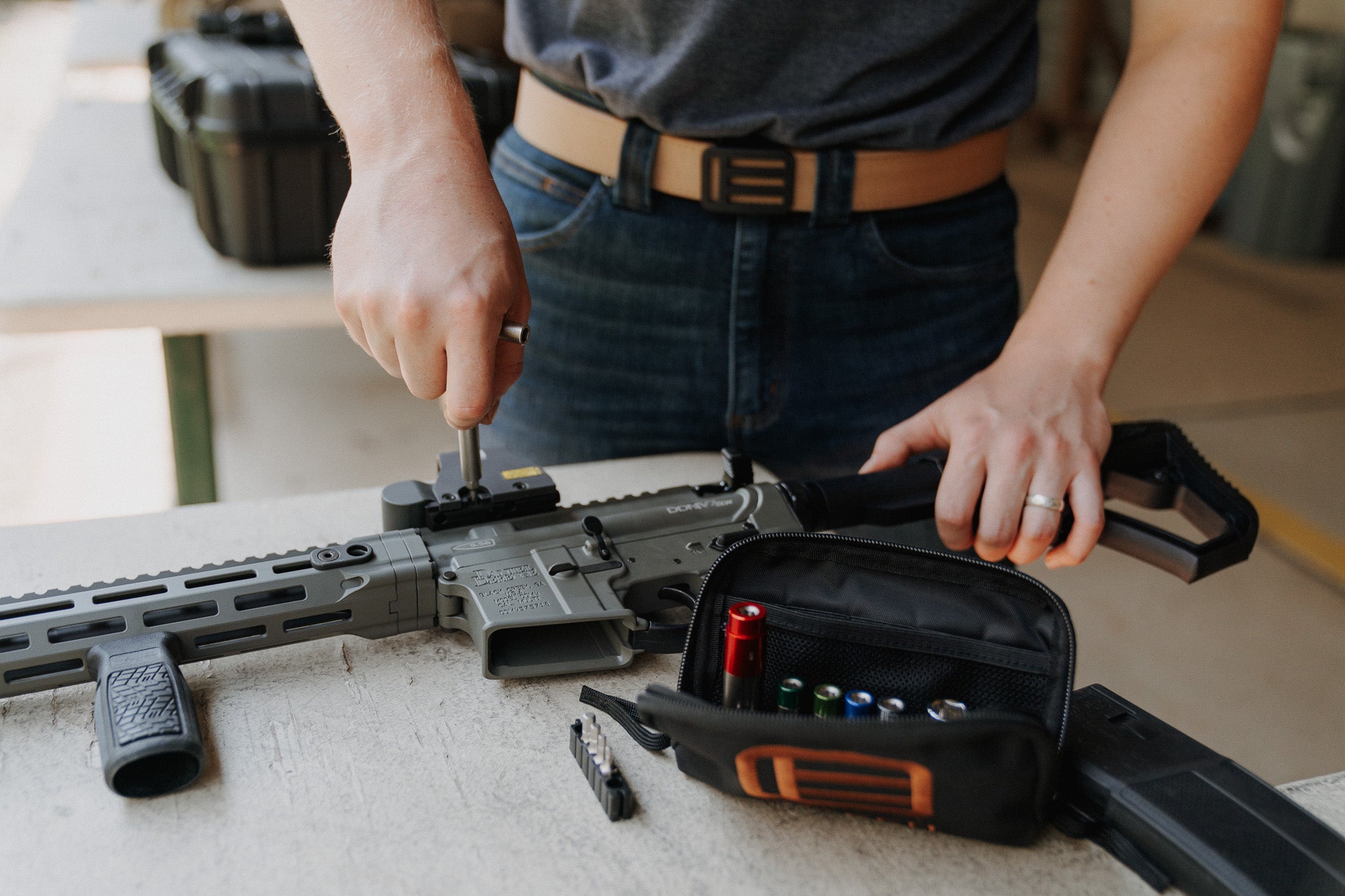 Person working on a rifle with fix it sticks tools and accessories on a table, wearing a tan EOTECH EDC belt