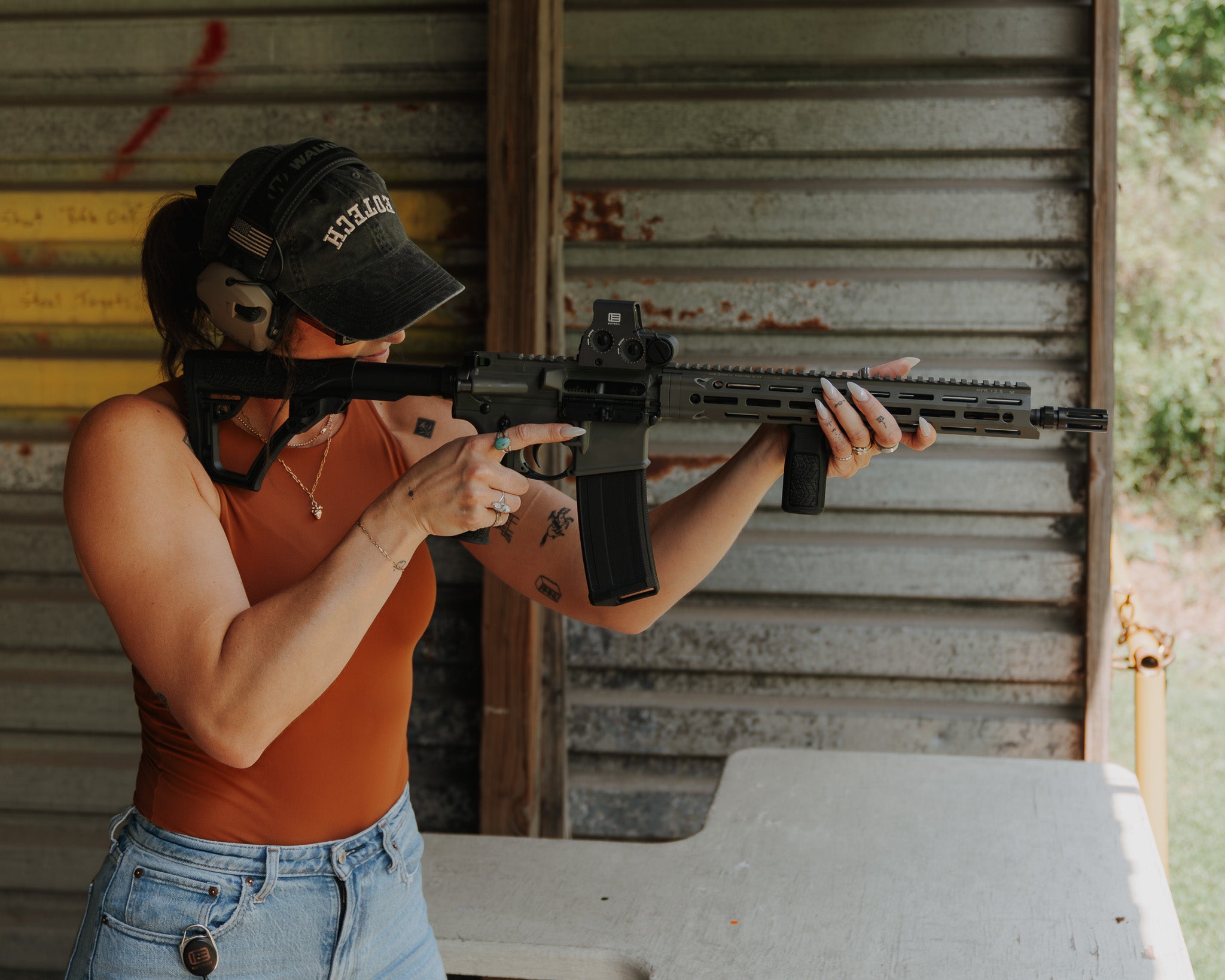 Person holding a rifle in front of a corrugated metal wall wearing black cap with EOTECH upside down