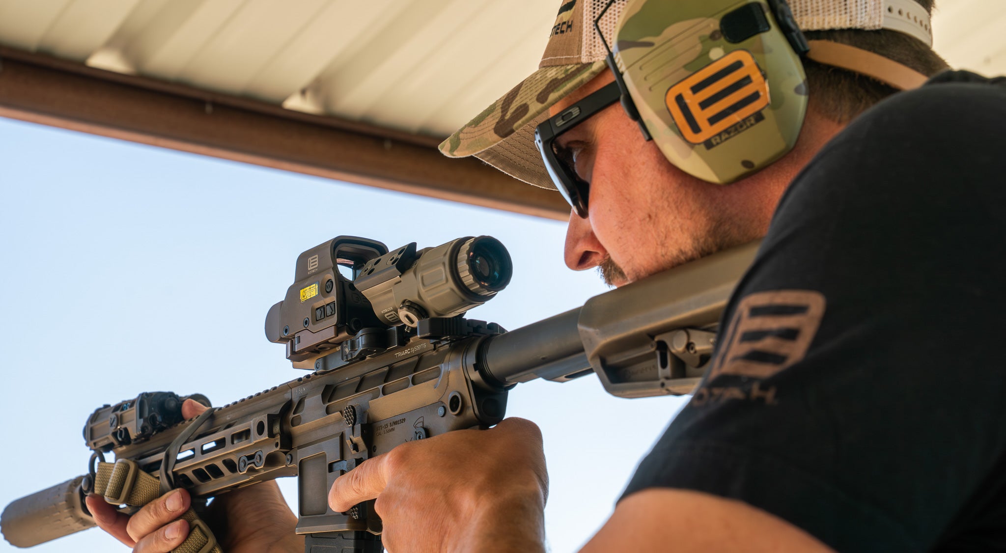 Person holding a rifle with a tan EOTECH HWS optic and magnifier, wearing EOTECH hearing protection and a hat, against a clear sky.