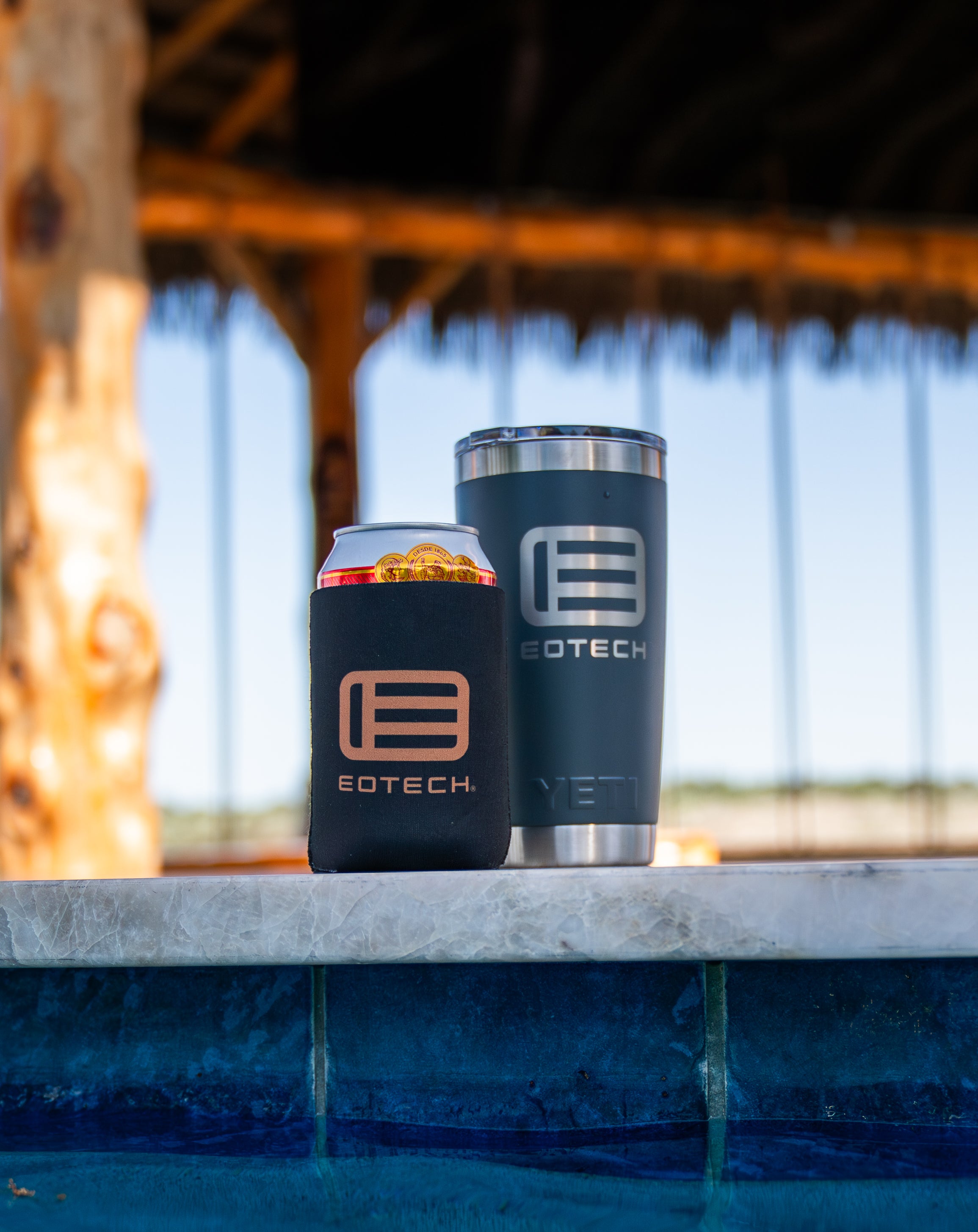 Eotech branded tumbler and can cooler on a ledge with a blurred natural background