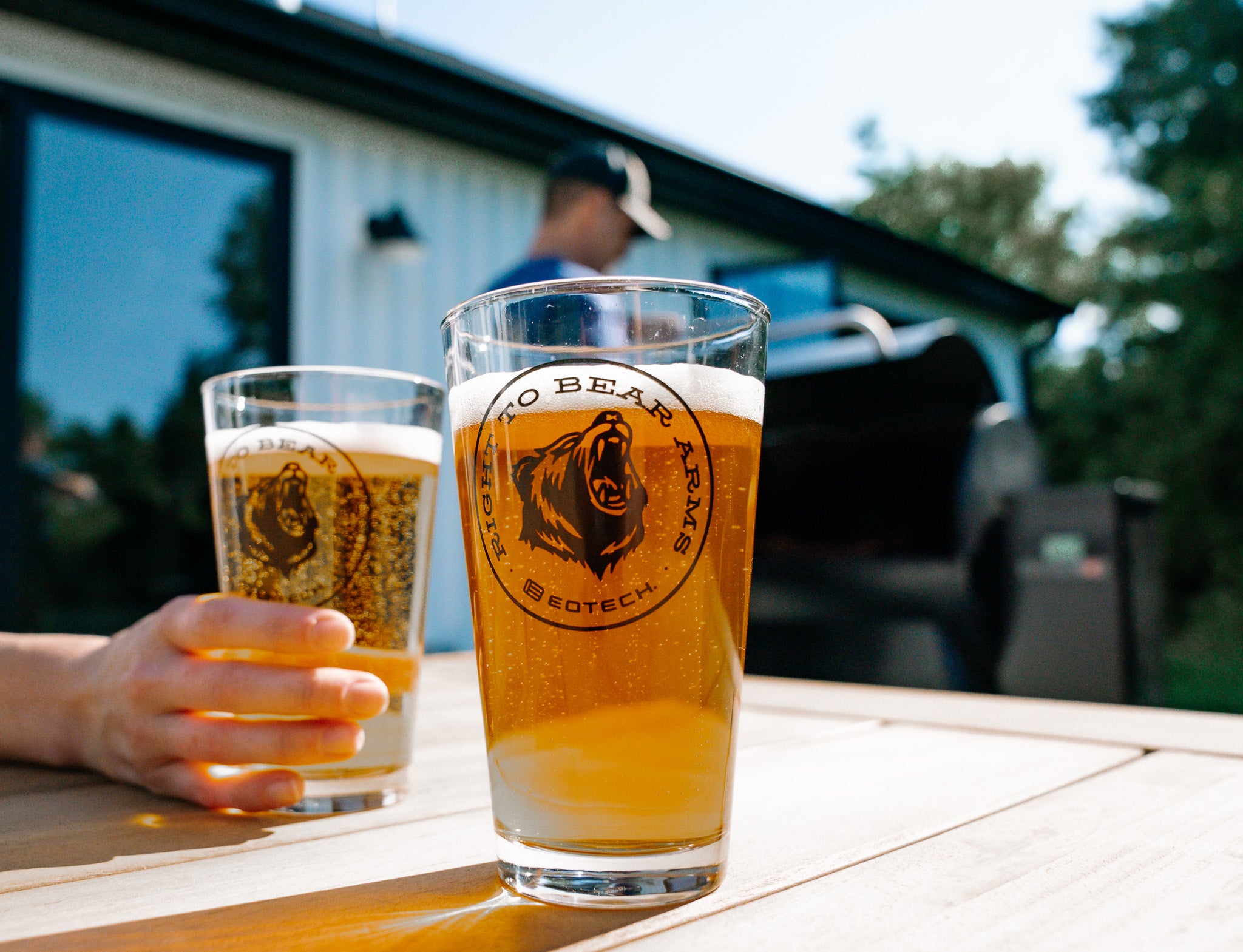 Two glasses of beer with a EOTECH logo on a table outdoors, with a person in the background.