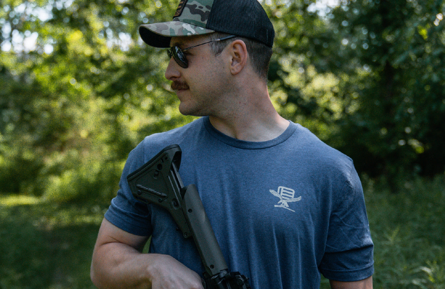 Man wearing a blue t-shirt with EOTECH logo, holding a rifle in a forest setting