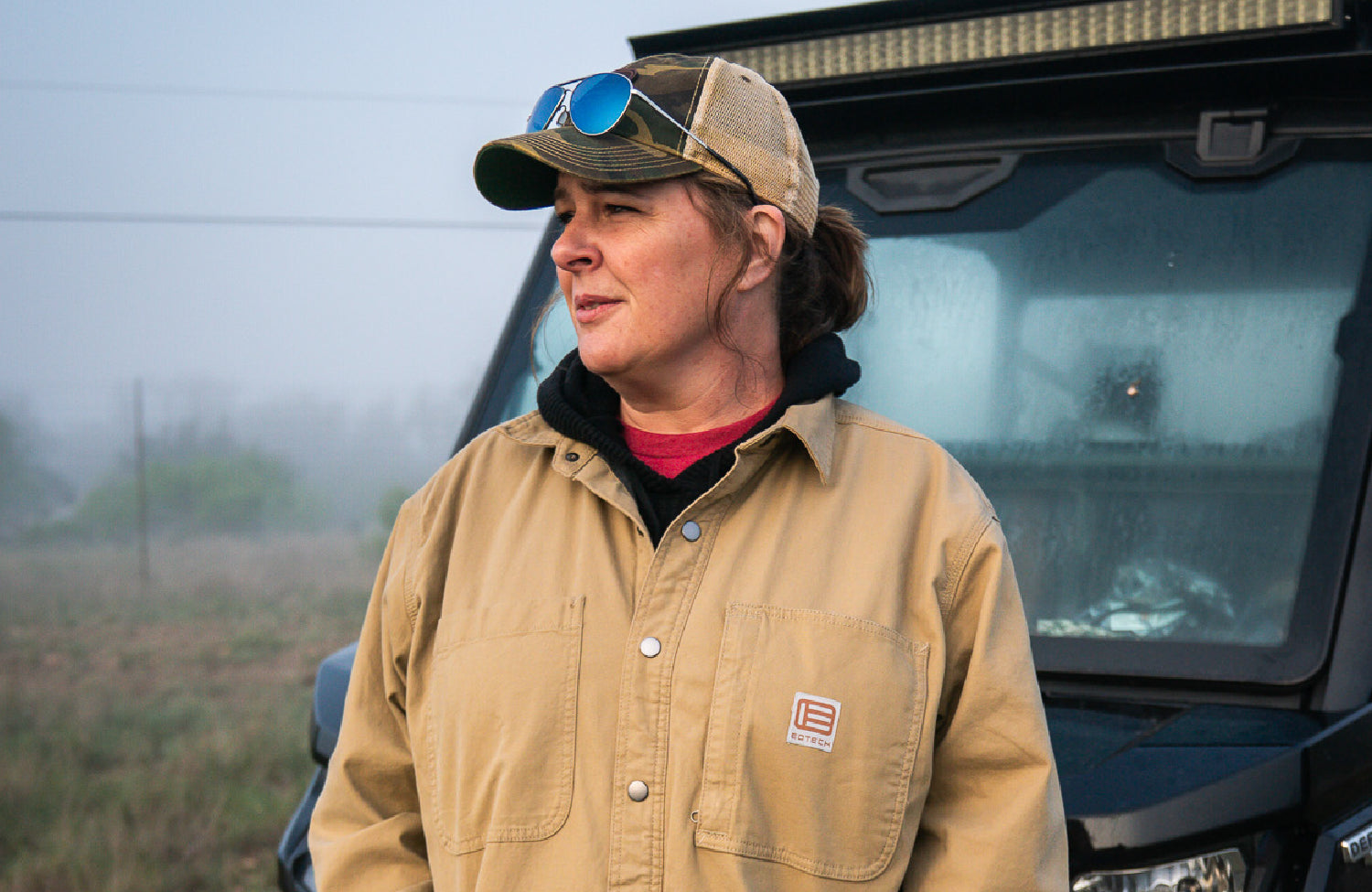 Woman wearing a tan EOTECH jacket and cap standing next to a vehicle in a field