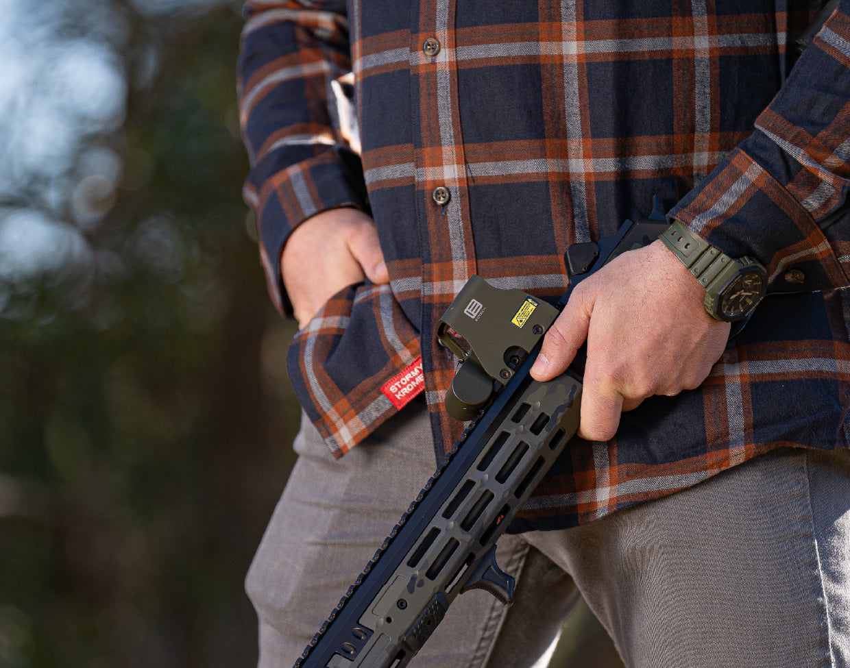 Person holding a rifle with a EOTECH HWS optic, wearing a plaid jacket and watch, outdoors.