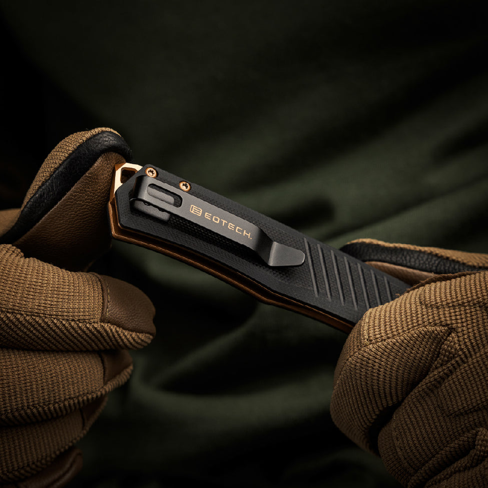 Black EOTECH pocket knife held between brown gloves with a dark background