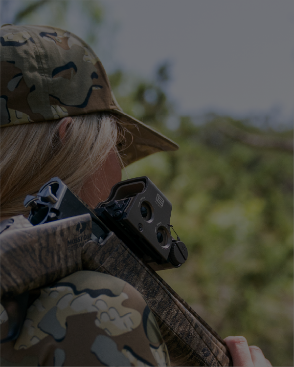 Female in camouflage clothing and hat holding a rifle with EOTECH HWS optic, outdoors.