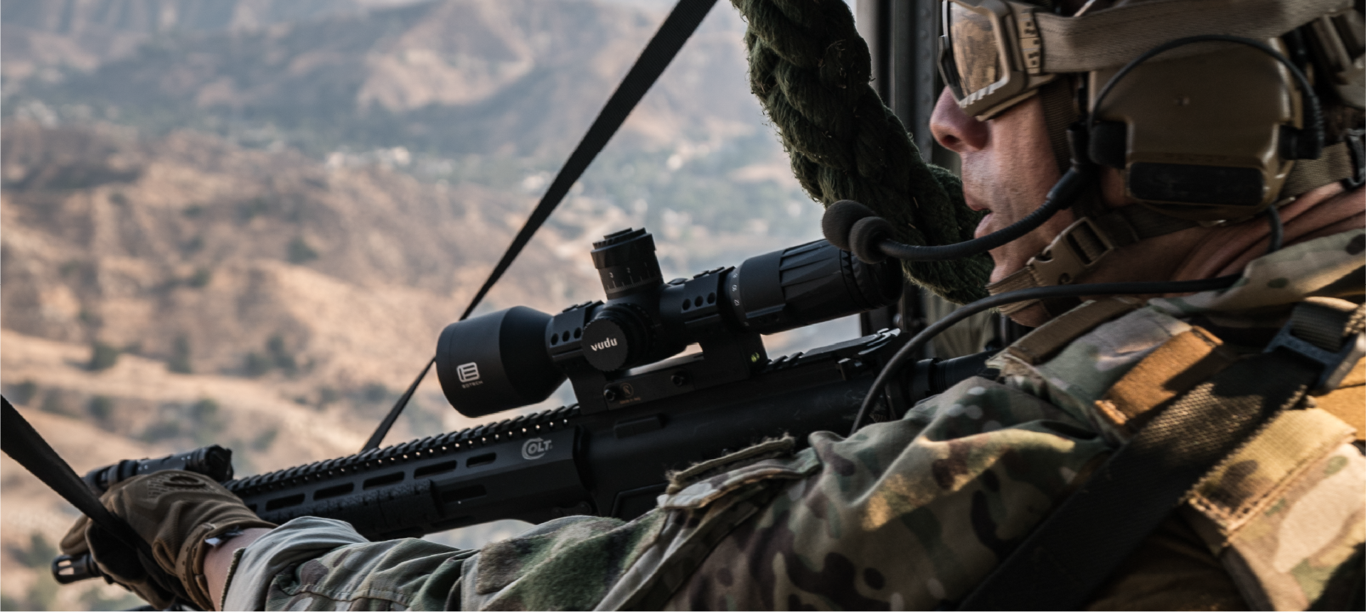 Soldier with rifle and EOTECH Vudu scope inside a helicopter with mountainous landscape below