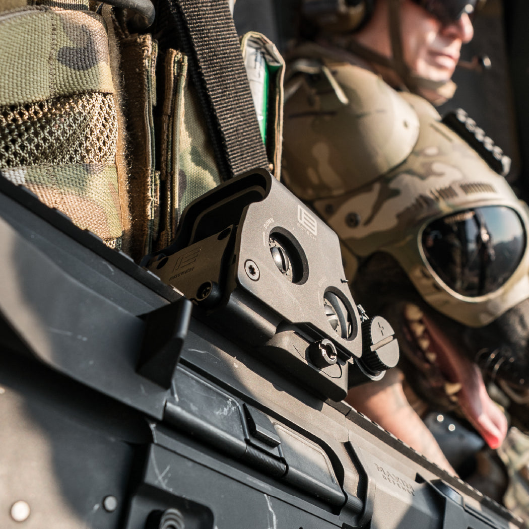 Close-up of a tactical rifle with tan EOTECH HWS optic with a military person and dog in camouflage gear in the background