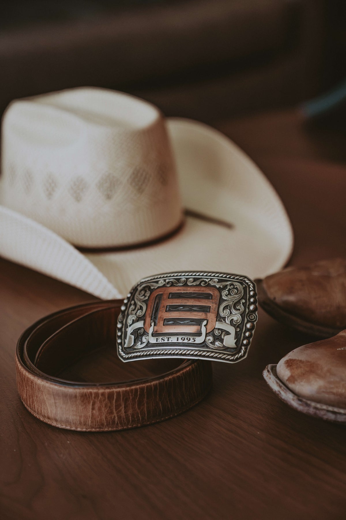 A western belt with a large engraved silver buckle featuring a copper-toned 'E' and 'EST. 1995' text, displayed on a wooden surface next to a white cowboy hat and brown leather boots.