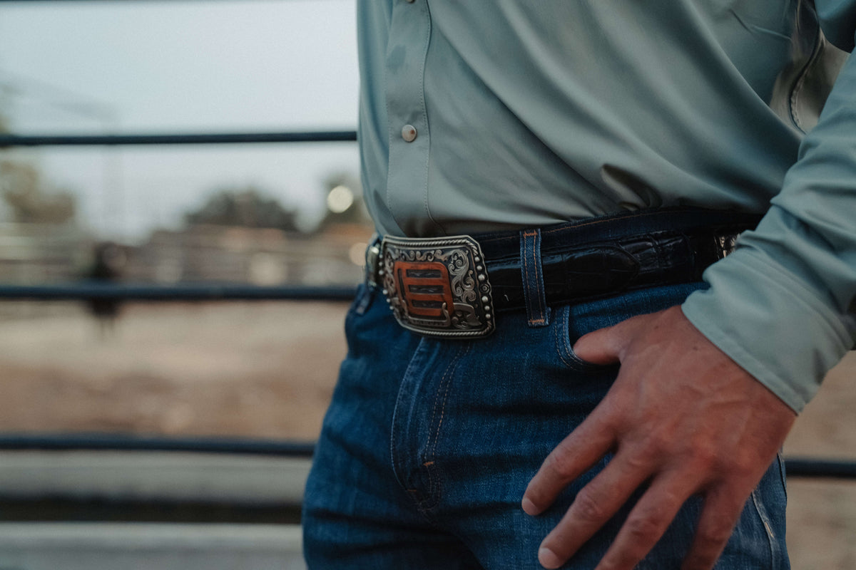 A close-up lifestyle shot of a man wearing a black leather belt with a large, ornate silver-toned western-style buckle featuring a red center panel, paired with a light blue button-down shirt and blue jeans.
