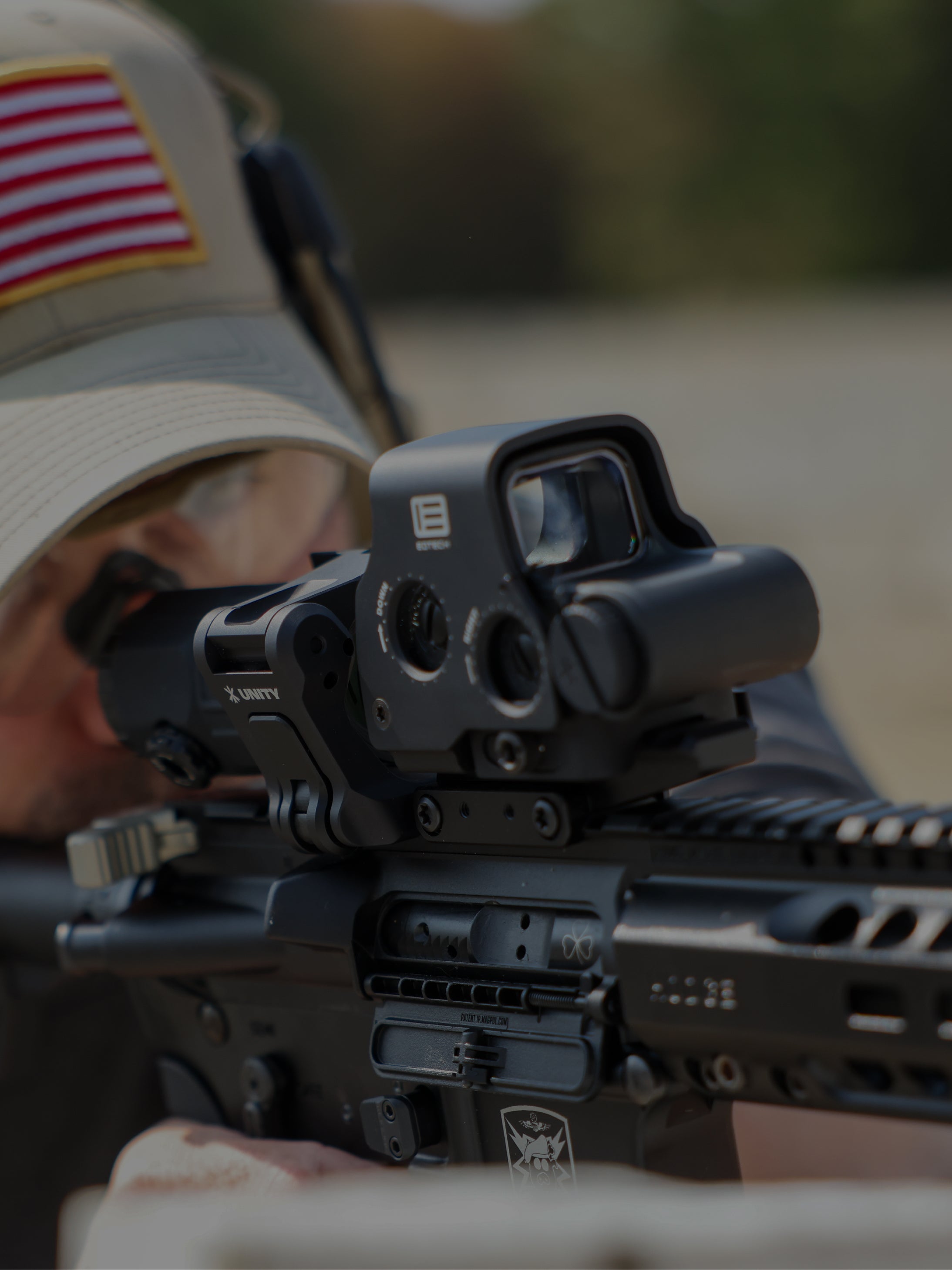 Close-up of a black EXPS3 with UNITY Mounts mounted on a rifle, with a person in a beige cap aiming the firearm outdoors.