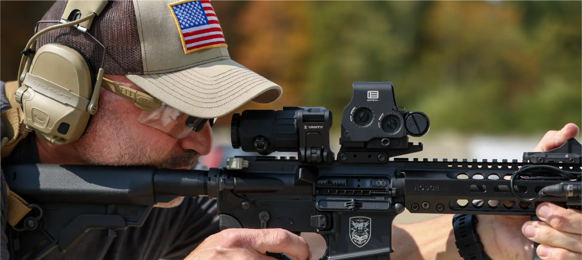 Person wearing tactical gear and holding a rifle with a HHS UNITY optic system, outdoors.