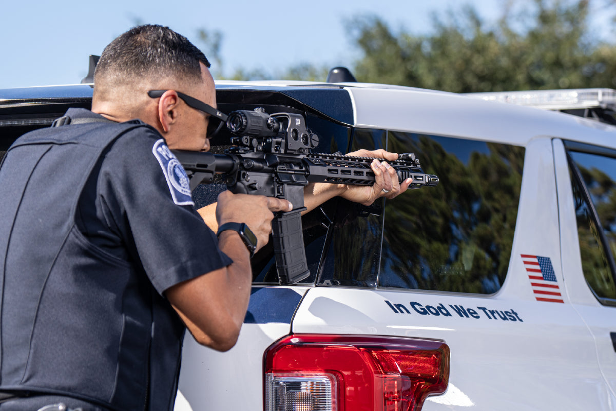 Law Enforcement Person aiming a rifle with black EOTECH HWS optic and magnifier in front of a police vehicle