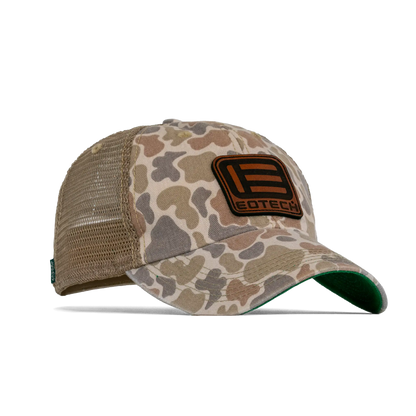 A duck camouflage trucker hat with tan mesh back panels and a brown leather patch on the front featuring the EOTECH logo from an angle.