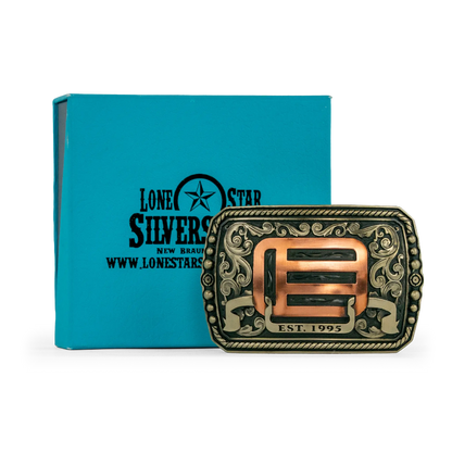 An ornate western-style belt buckle with gold-toned metal edges, and copper-toned scrollwork and geometric details, shown alongside its box on a white background.