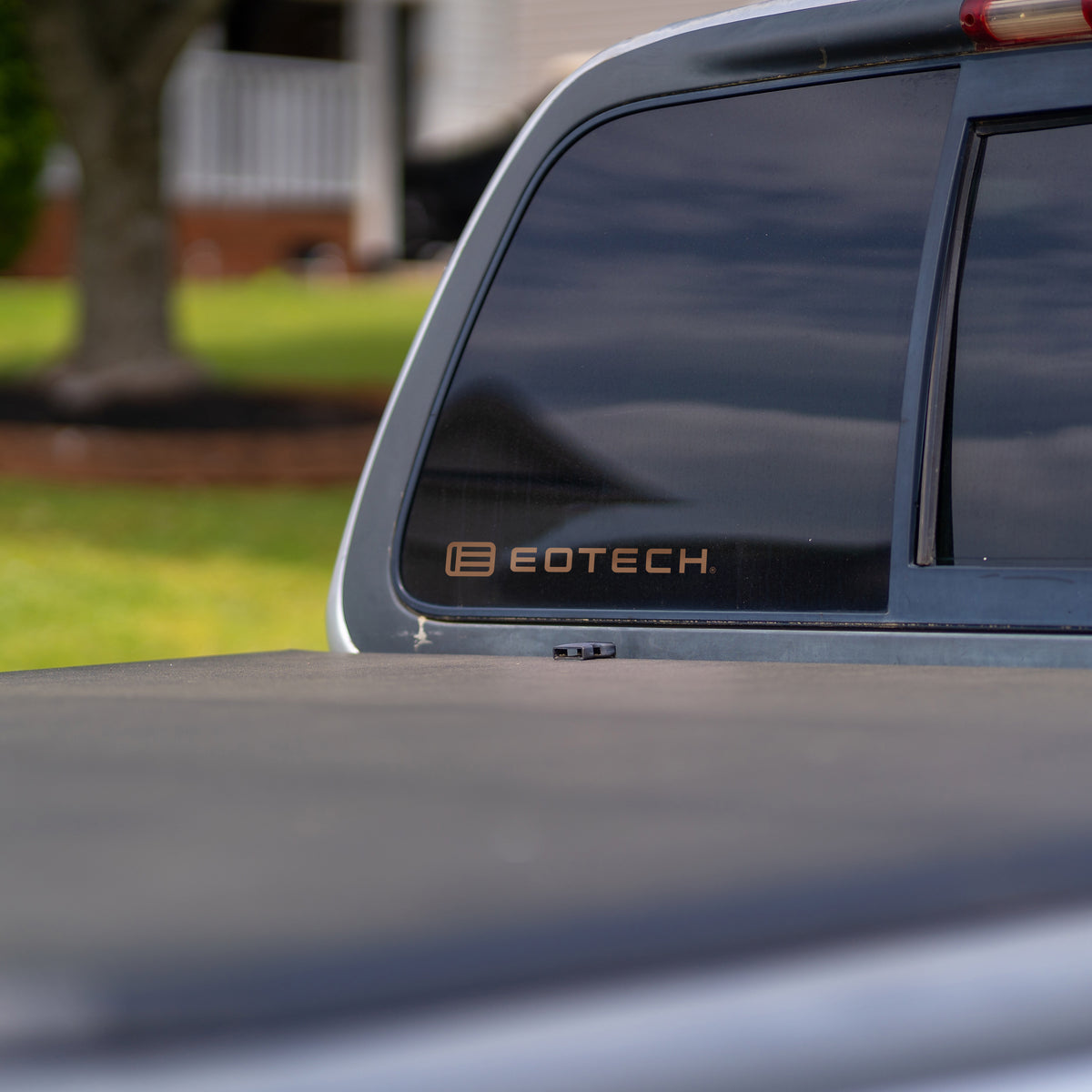 Close-up of the rear side window of a dark pickup truck with an EOTECH decal on the window, with a blurred outdoor background showing grass and a house