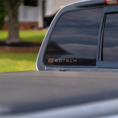 Close-up of the rear side window of a dark pickup truck with an EOTECH decal on the window, with a blurred outdoor background showing grass and a house