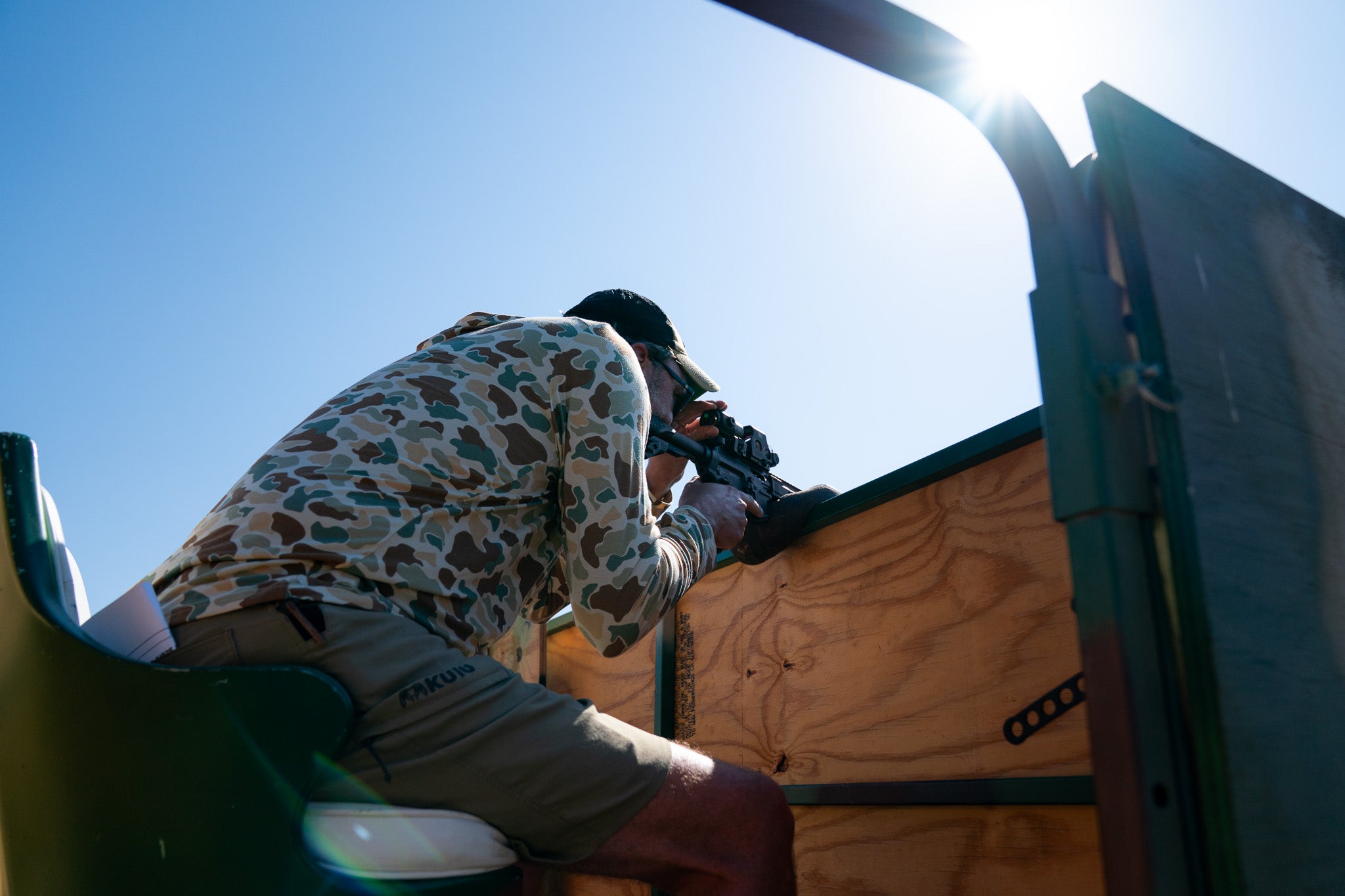 Person in camouflage Free Fly hoodie and hat shooting on a wooden structure with a clear blue sky.