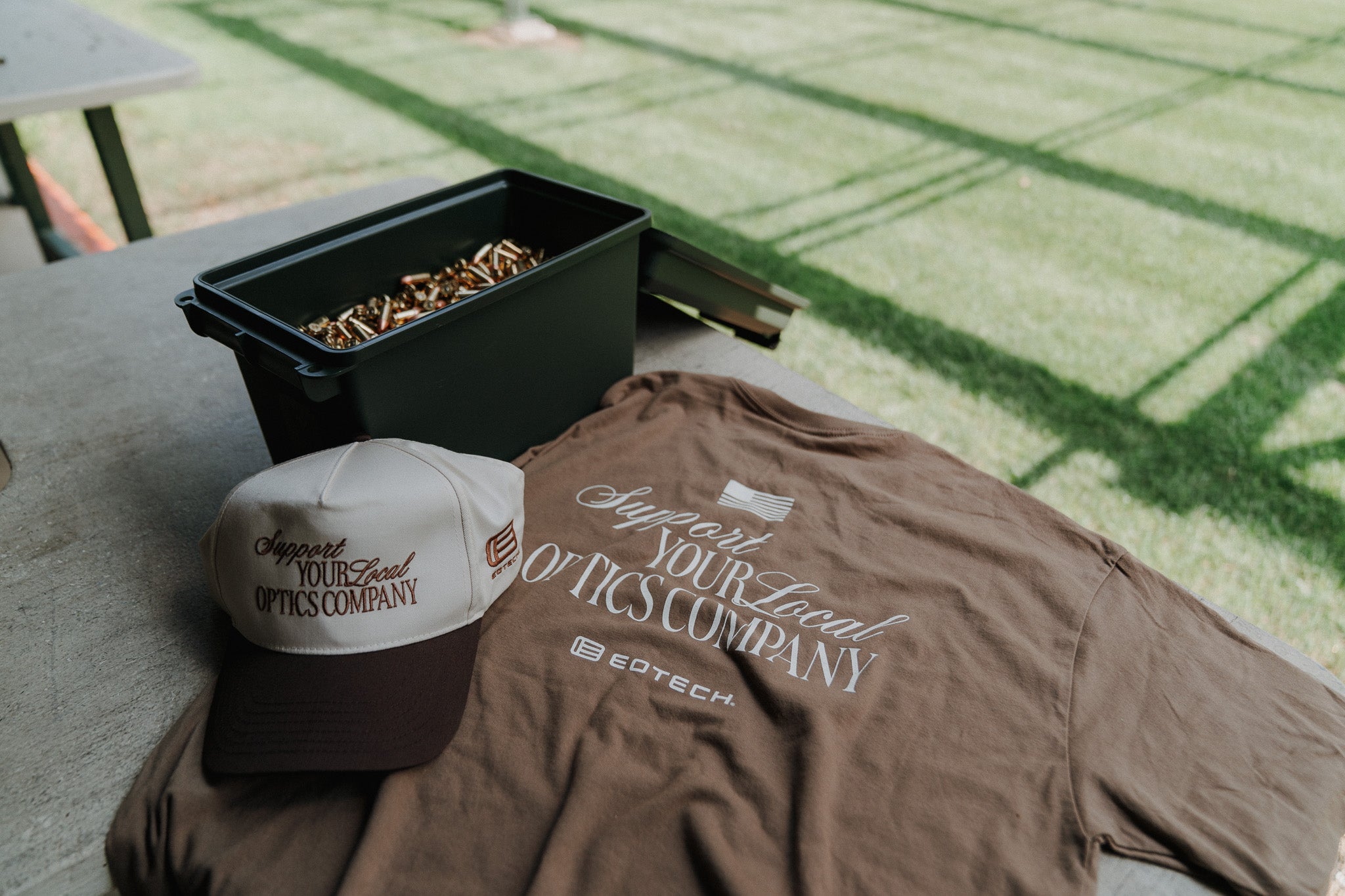 Brown Support you local optics company t-shirt and cap on a table at a shooting range