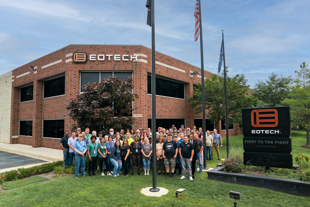 A large group of people posing outdoors in front of an Eotech brick building, with a black Eotech sign on the lawn and flagpoles in the foreground.