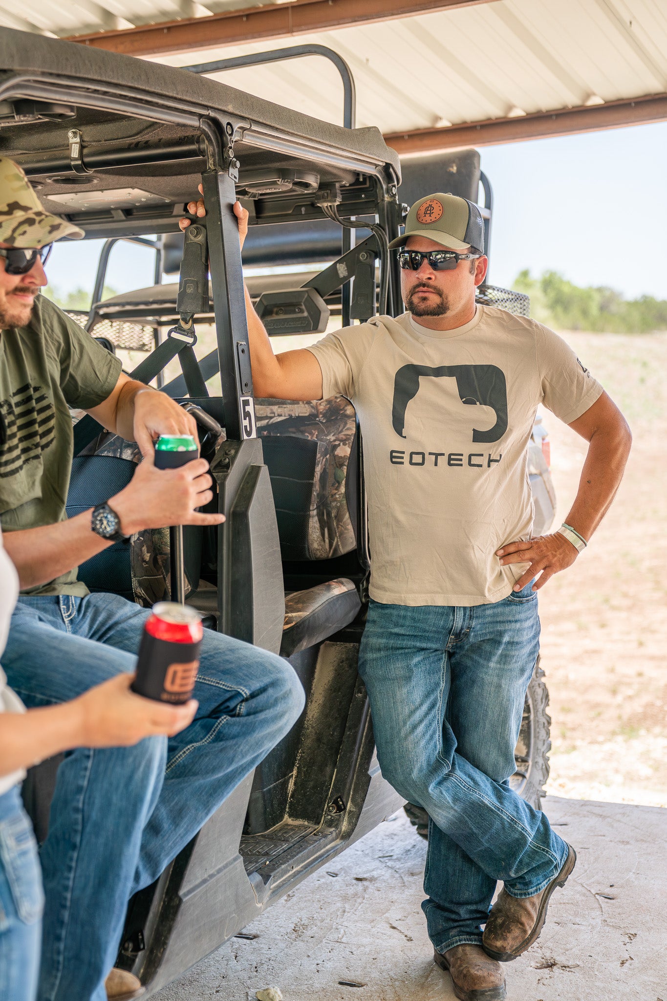 Two men in a vehicle, one wearing a EOTECH shirt, with a desert landscape in the background.