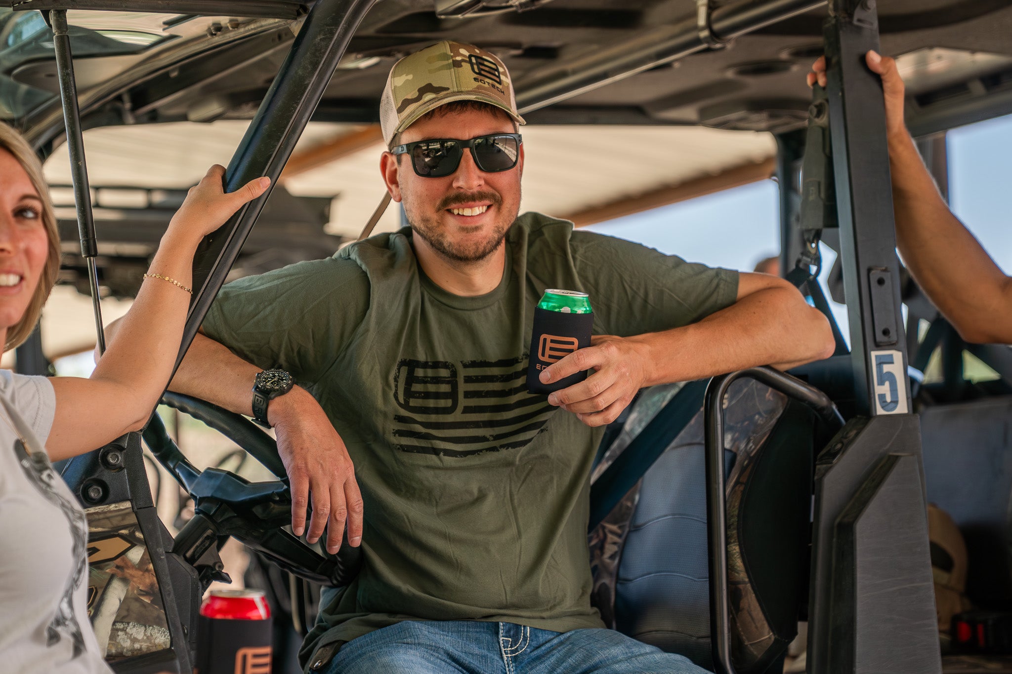 Man with EOTECH shirt sitting in an off-road vehicle holding a beer in EOTECH kookie, with a woman beside him.