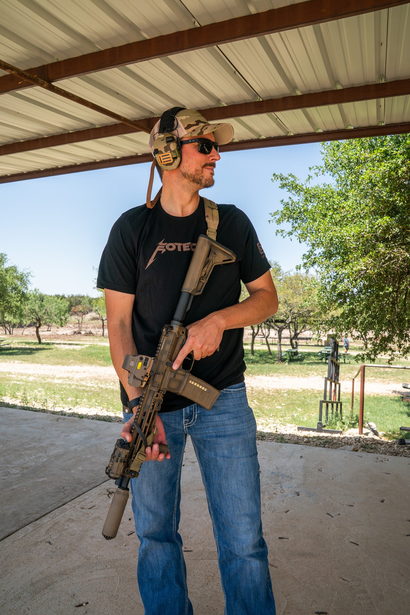 Man holding a rifle with tan EOTECH HWS optic under a covered outdoor area with trees in the background