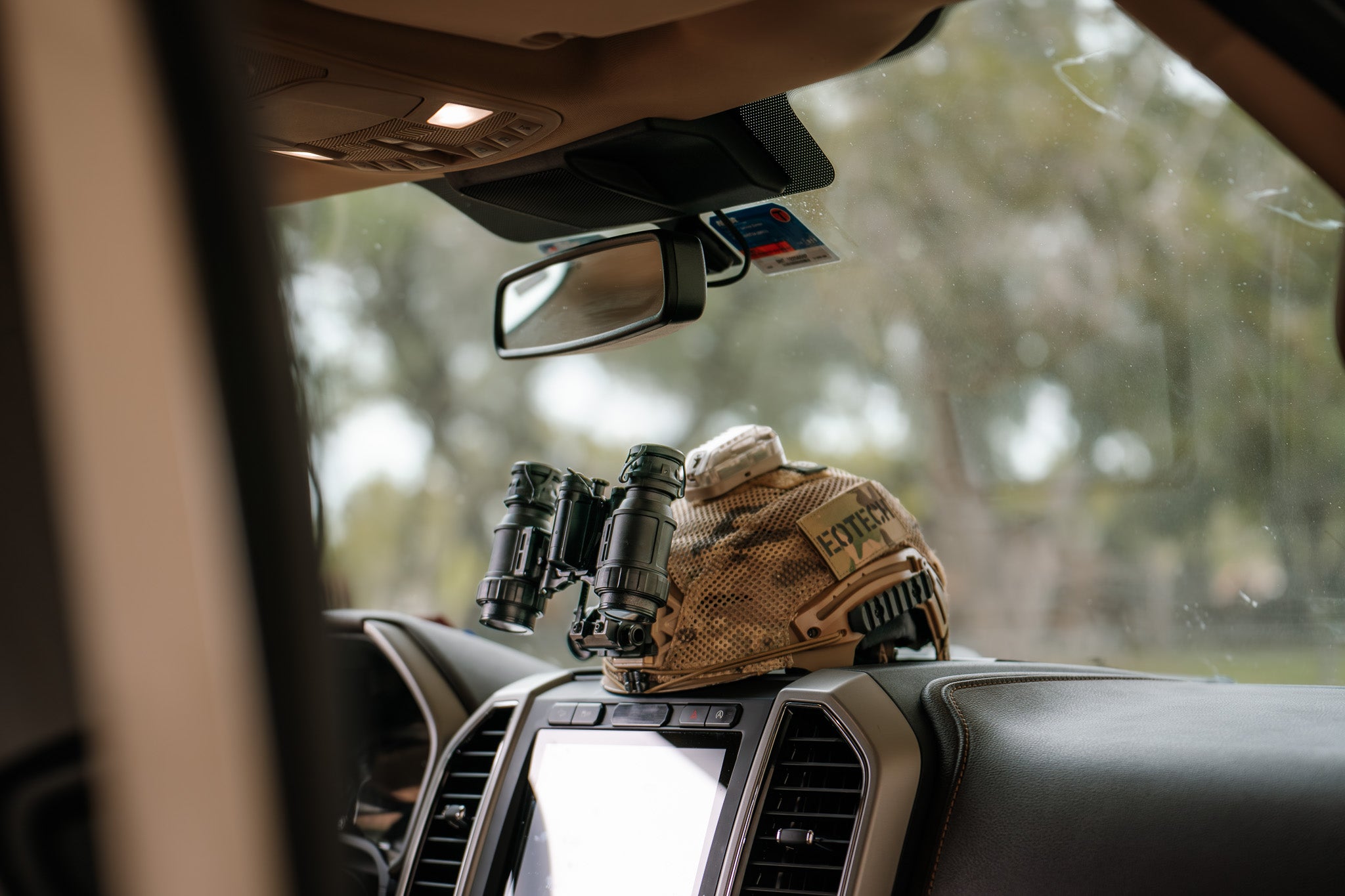 Camouflage-patterned helmet and EOTECH binoculars on a car dashboard with a blurred outdoor background
