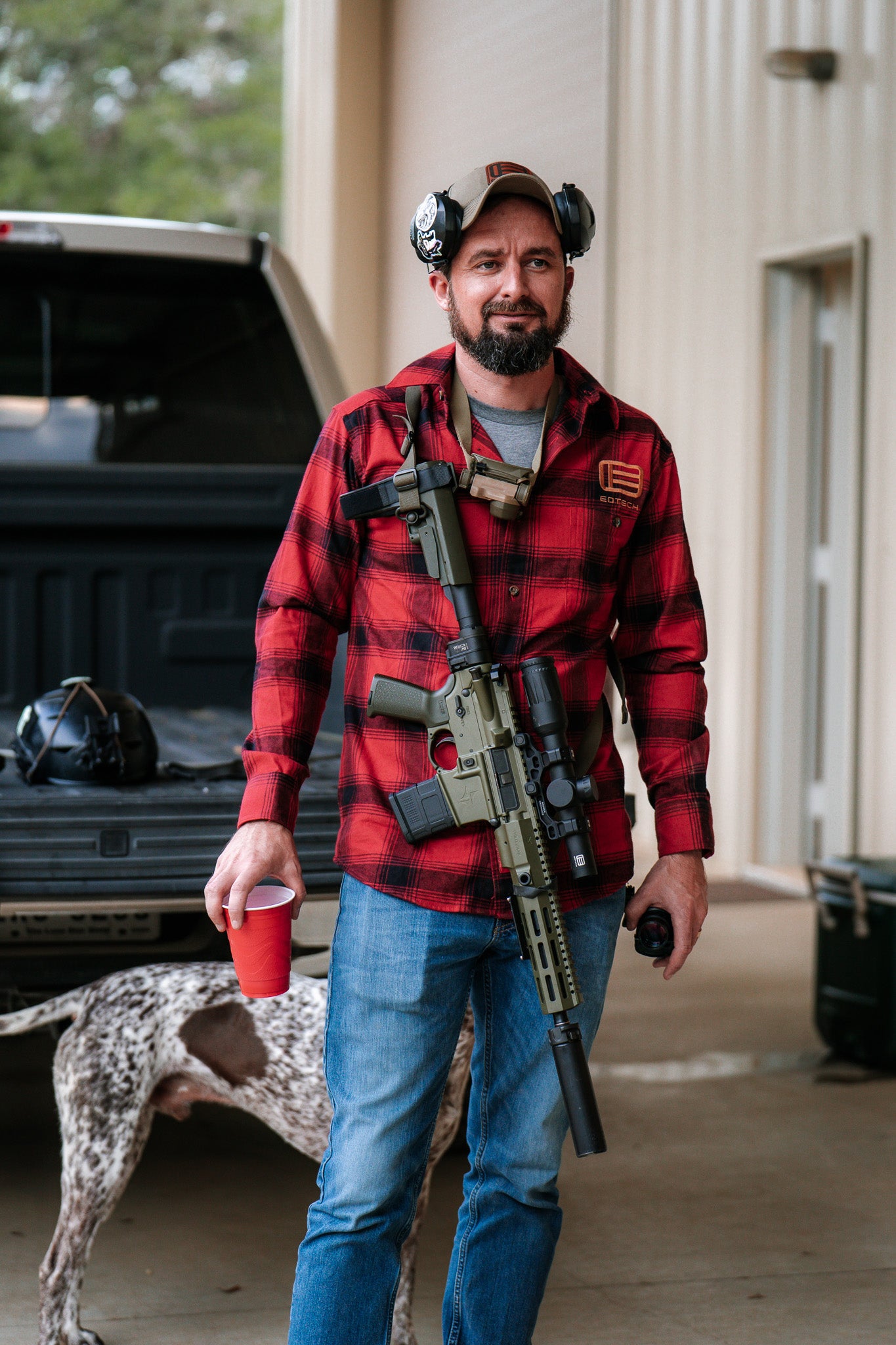 Man in red plaid shirt with EOTECH logo holding a rifle with Vudu optic, standing next to a dog with a building in the background