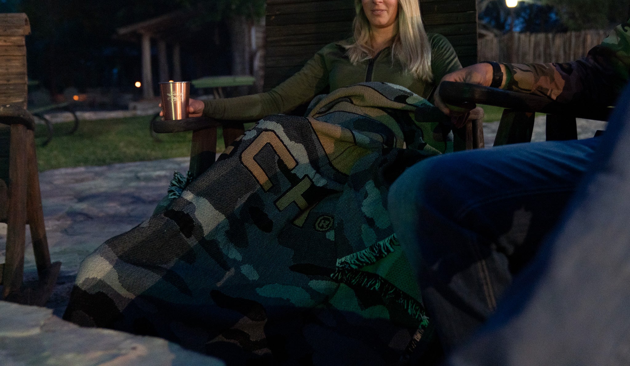 Woman sitting outdoors at night with a EOTECH blanket draped over her, holding a drink.