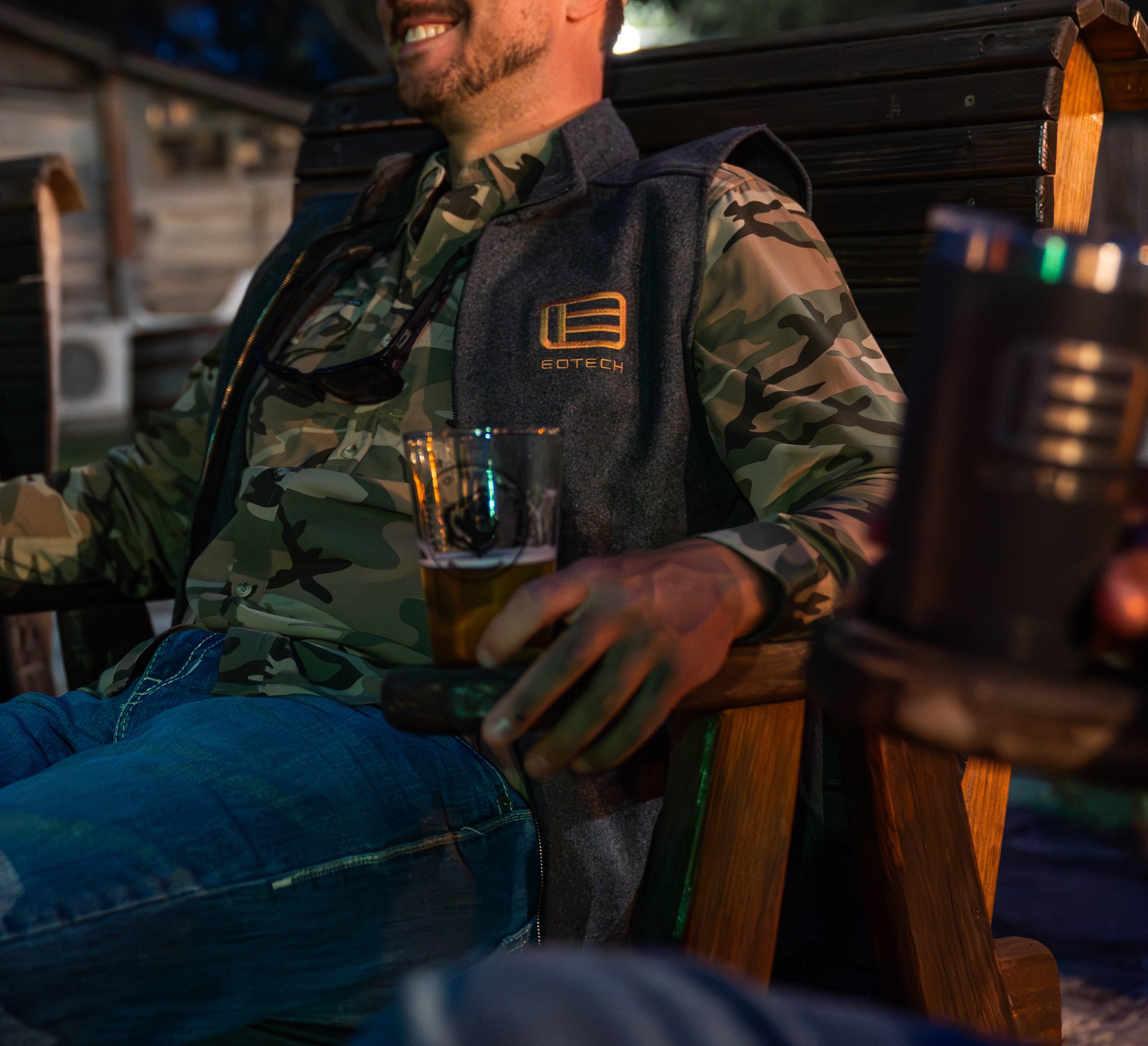 Man in camouflage jacket holding a EOTECH glass, sitting outdoors with a blurred background