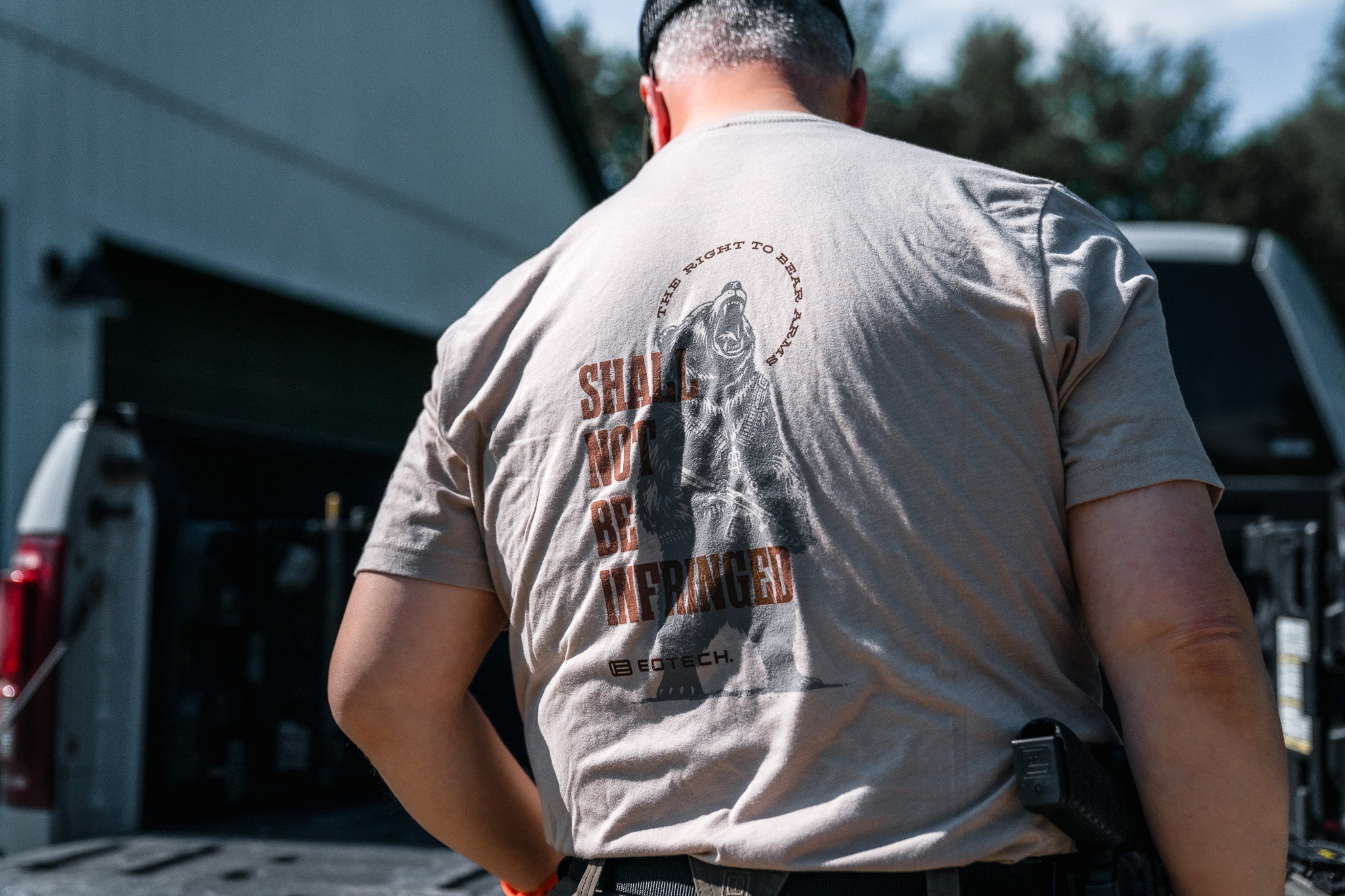 Person wearing a beige t-shirt with Shall Not Be Infringed text and graphics on the back, standing outdoors.
