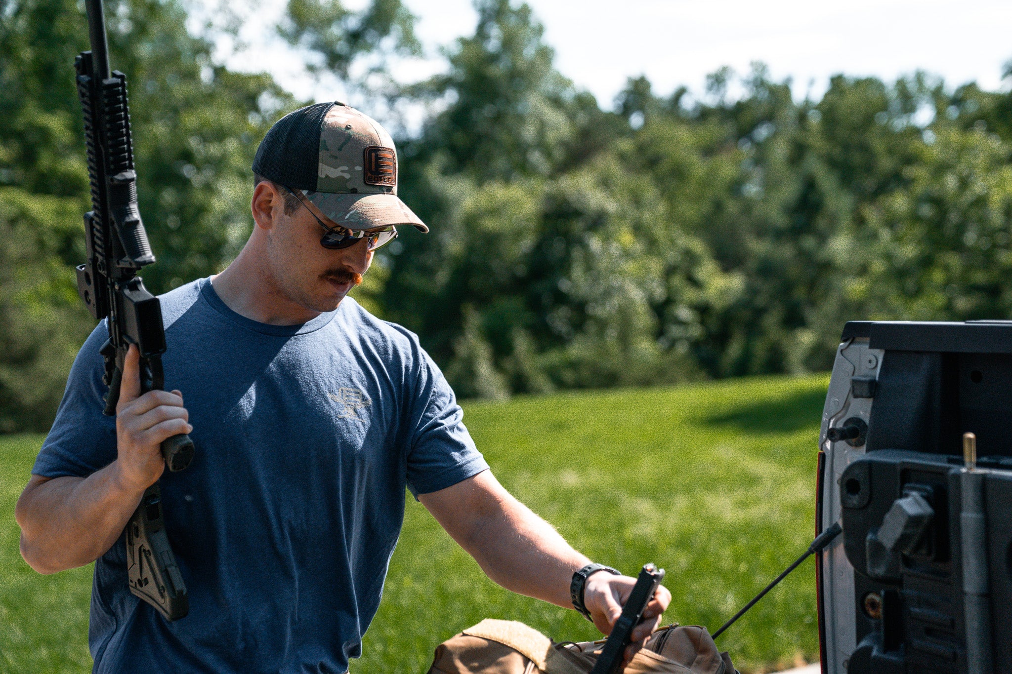 Man holding a rifle with EOTECH HWS optic and a gun case outdoors with trees in the background