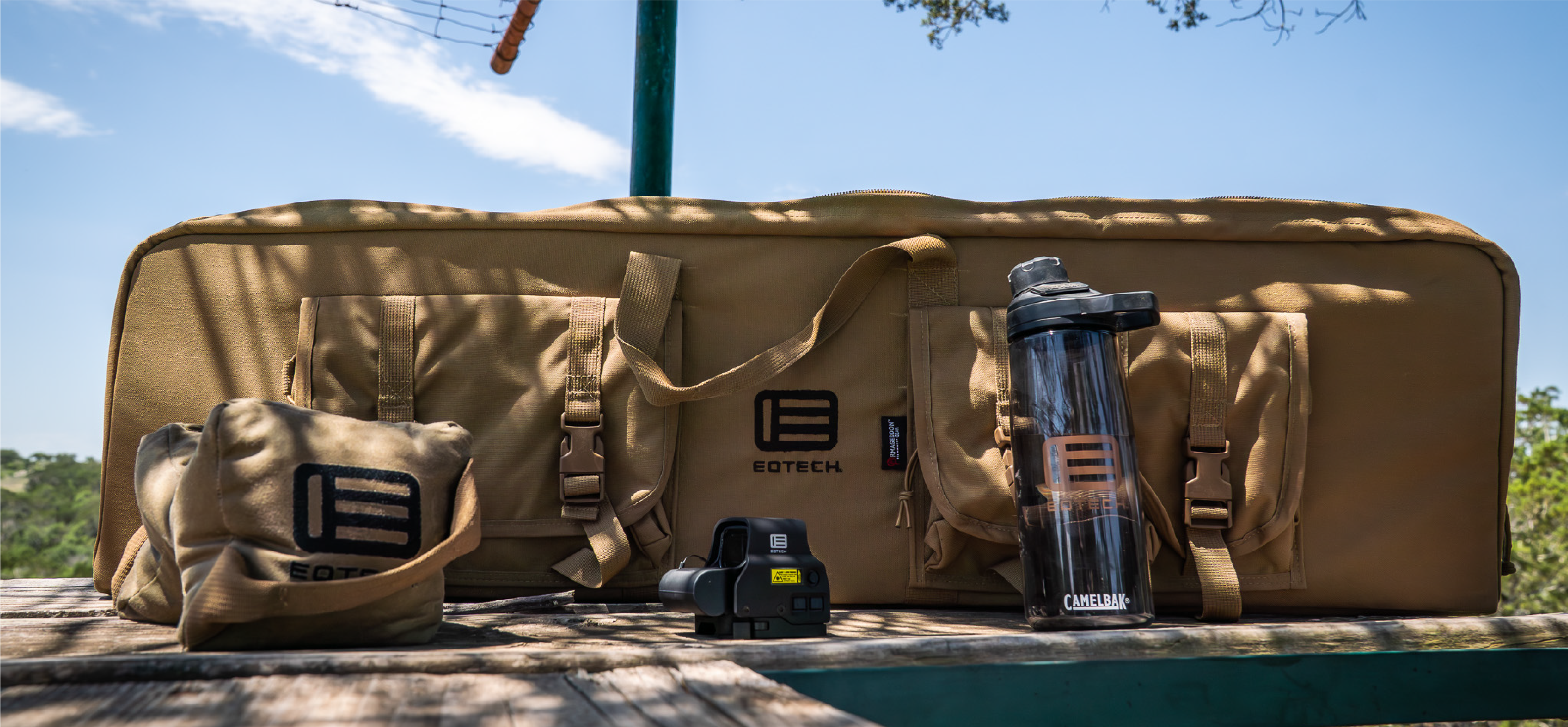 Coyote brown gun case with EOTECH logo, HWS optic, water bottle and shooting bag on a wooden surface outdoors.