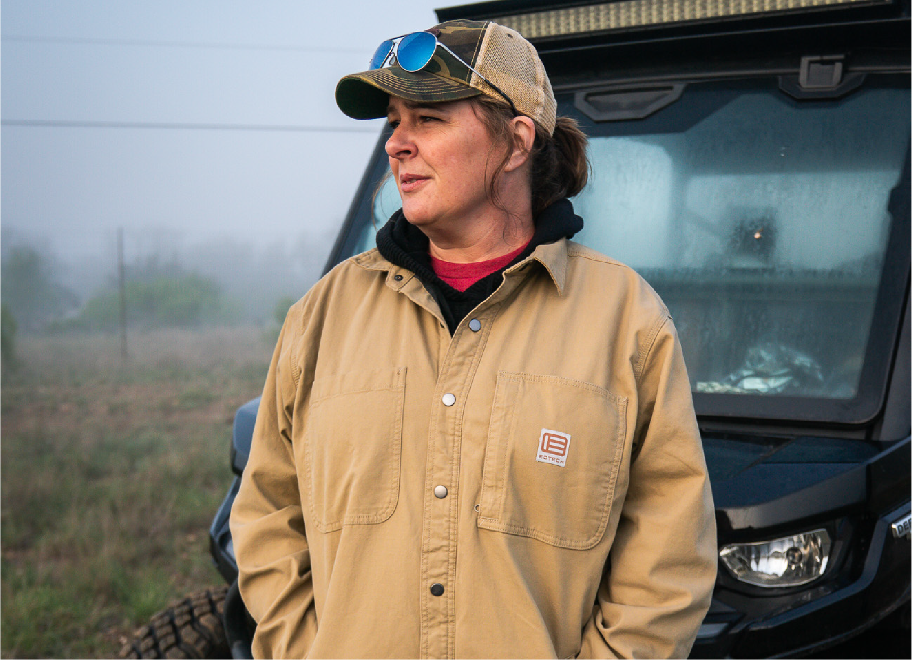 Woman wearing a EOTECH tan jacket and cap standing next to a vehicle in a field