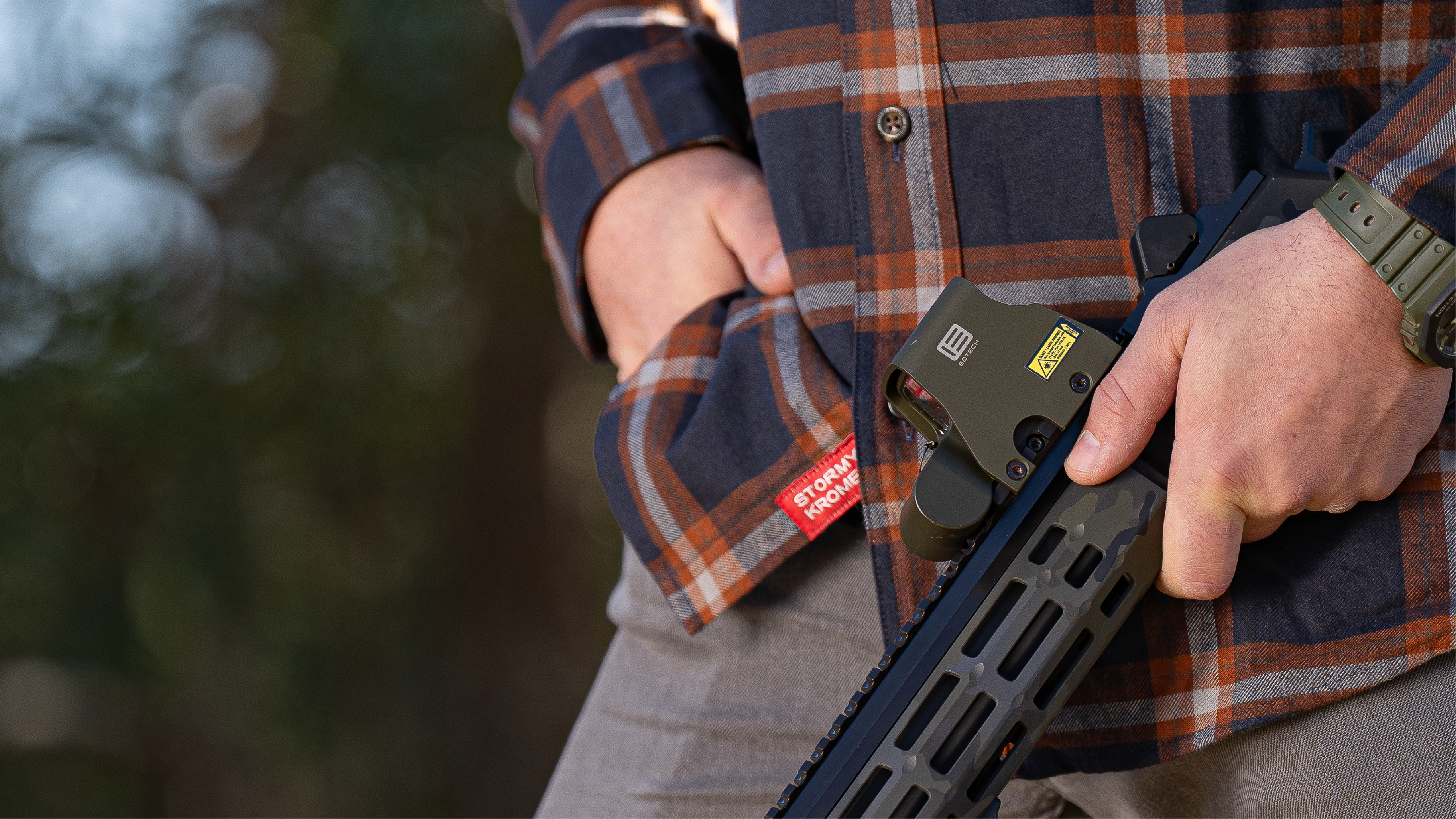 Person holding a rifle with a EOTECH HWS optic, wearing a plaid shirt, in an outdoor setting.