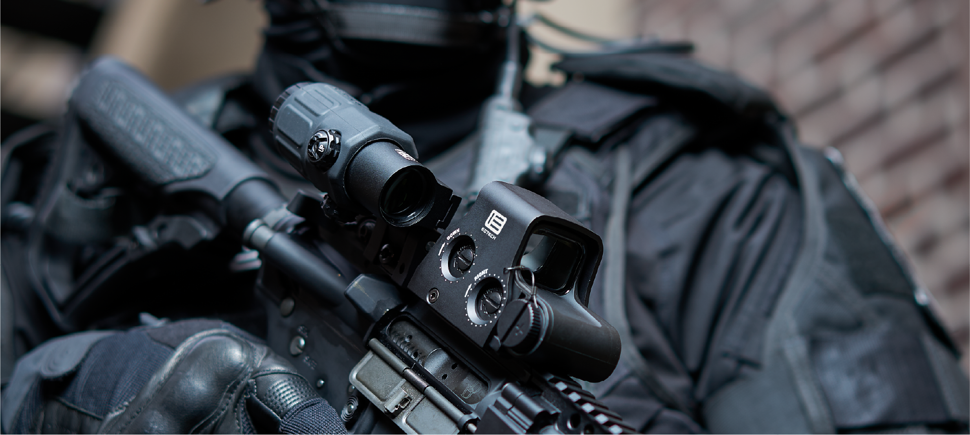 Person in tactical gear holding a rifle with a EOTECH HWS optic and magnifier, blurred background