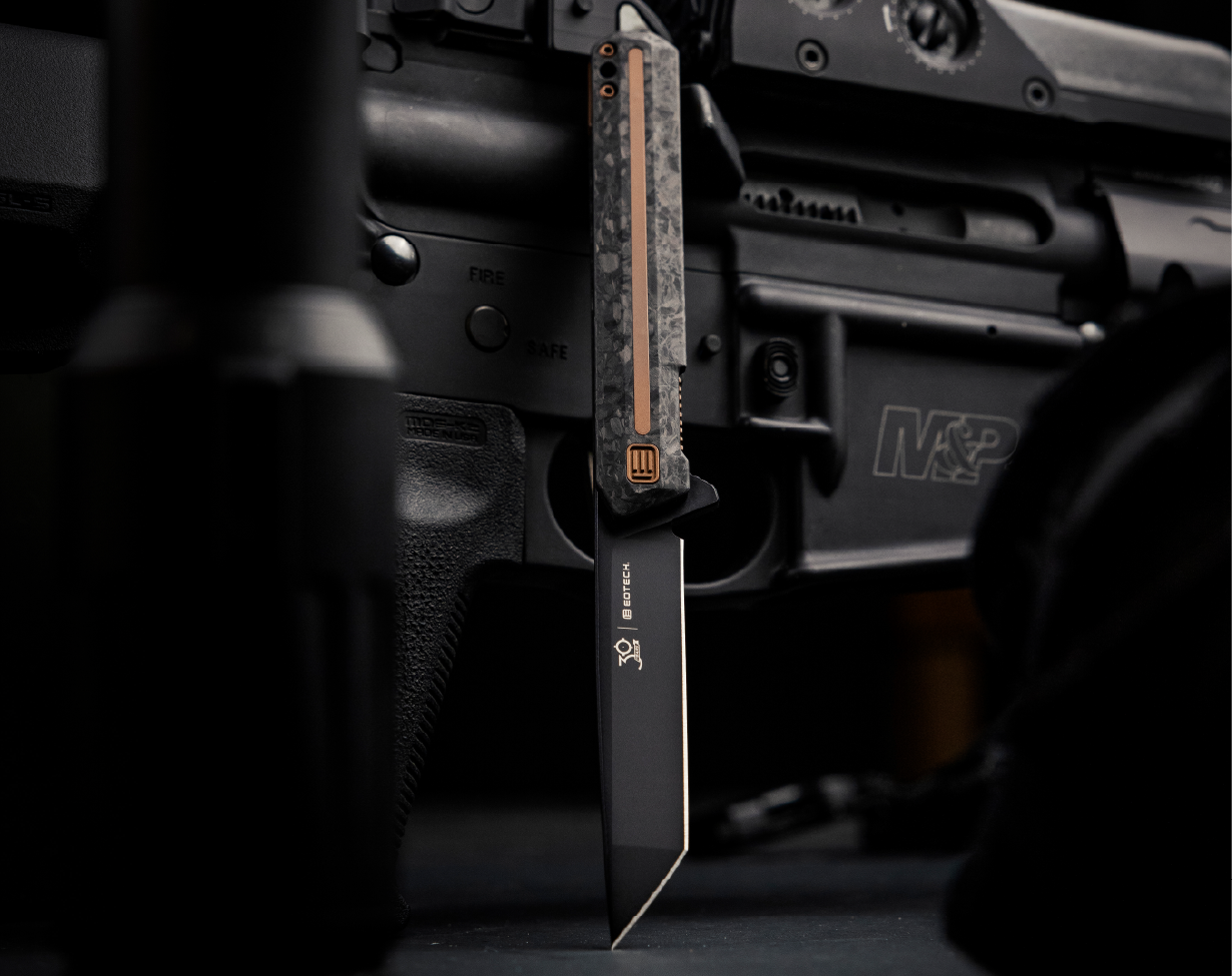 EOTECH Folding knife with a black blade and copper and black handle on a blurred background of a firearm.