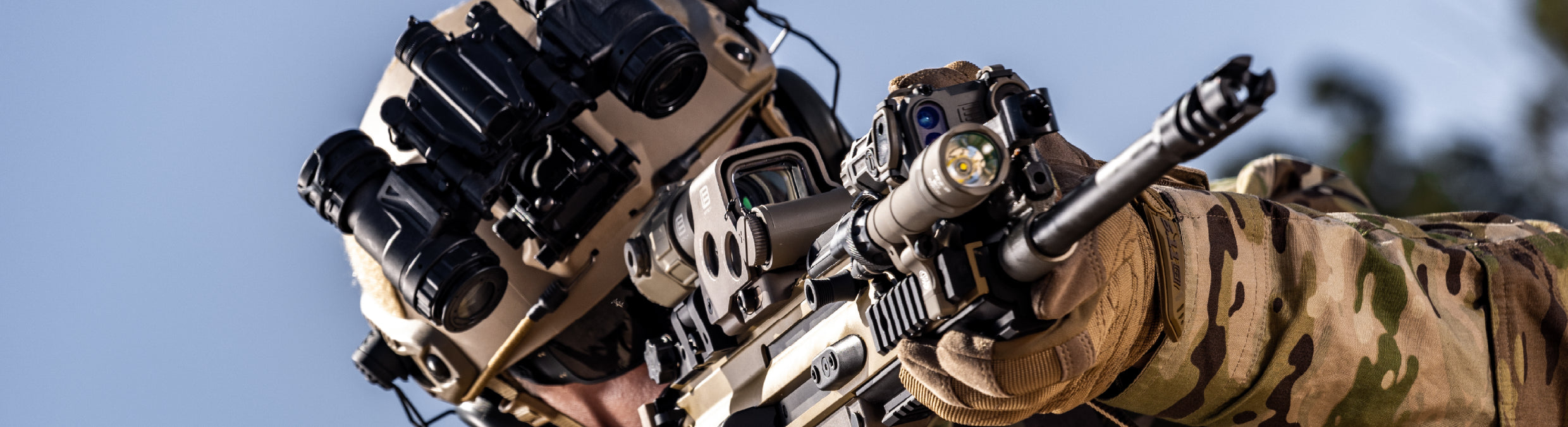 Person wearing a tactical helmet EOTECH binos and holding a rifle with tan HWS, Magnifier and OGL with a clear blue sky background