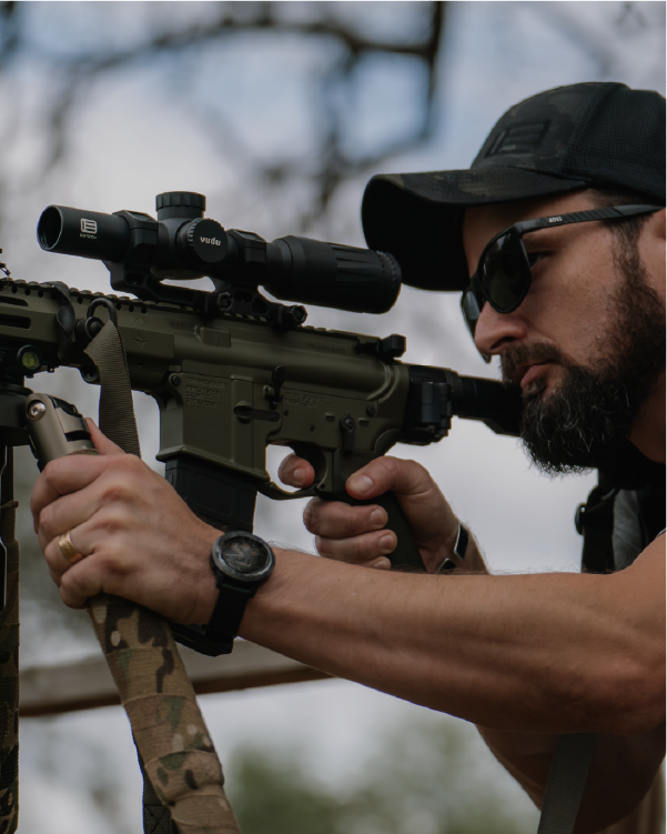 Man holding a rifle with a EOTECH Vudu scope, wearing sunglasses and a cap, outdoors.