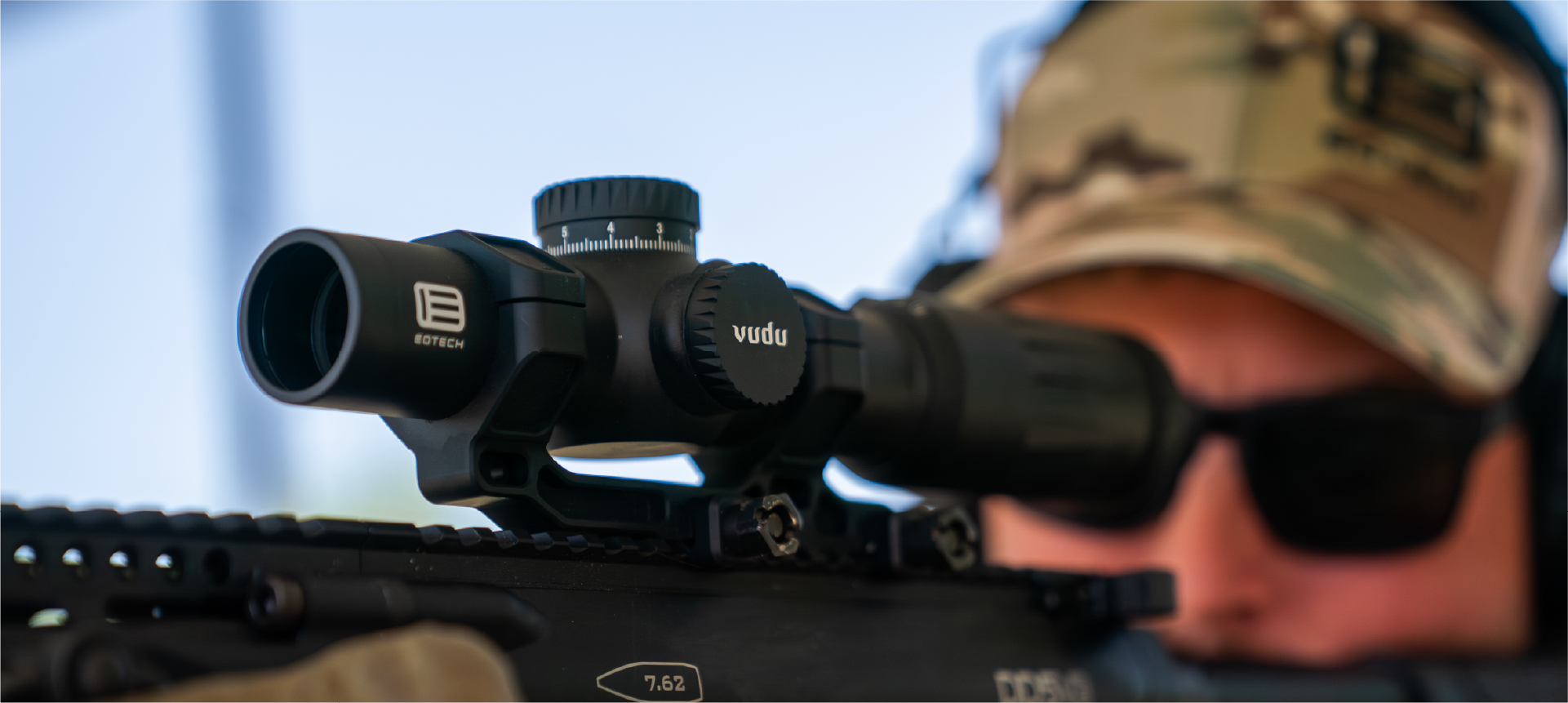 Person wearing a cap and sunglasses with a rifle equipped with a EOTECH Vudu black scope, blurred background