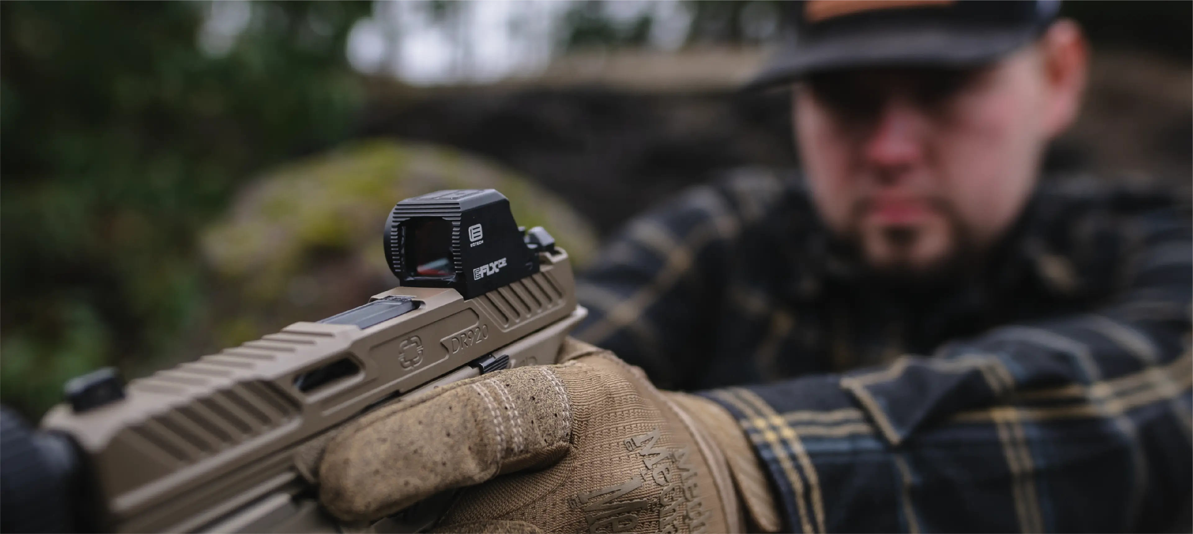 Person holding a pistol with an EFLX optic, wearing a plaid jacket and cap, outdoors.