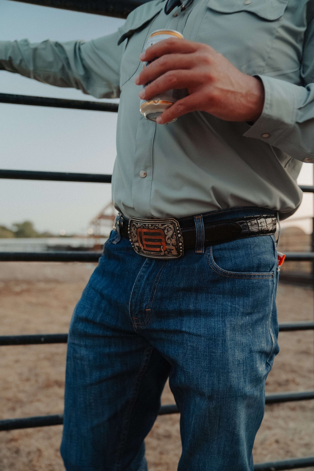 A man wearing a light sage green western-style button-up shirt, blue jeans, and a black leather belt with a large decorative silver buckle, holding a drink in an outdoor setting.