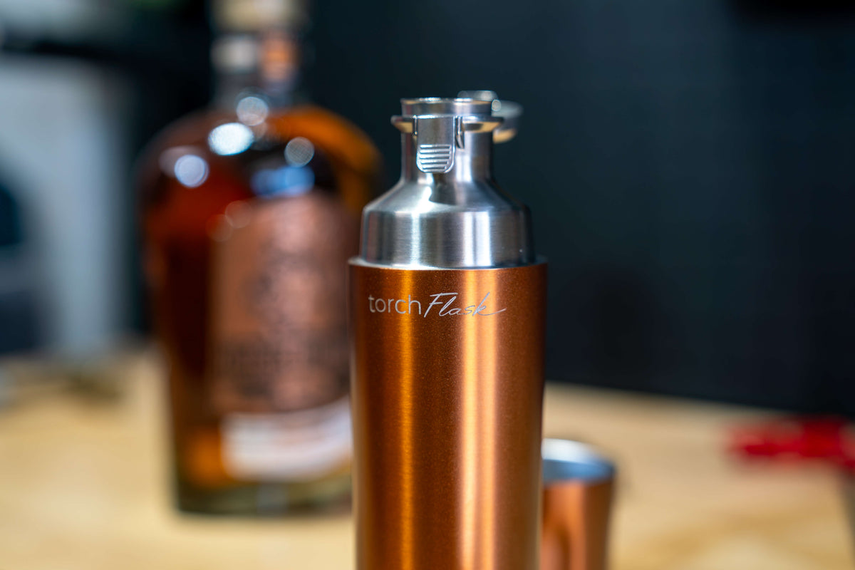 A copper-colored EOTECH Flash with a silver stainless steel top, featuring the 'torch Flash' logo, shown in a shallow depth-of-field setting with a blurred amber bottle in the background.