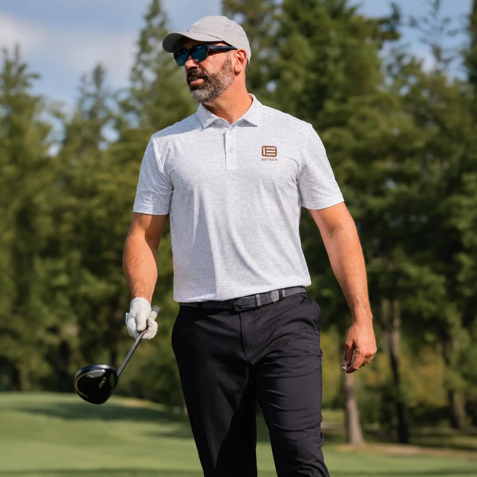 A man wearing a light EOTECH polo shirt with a small copper EOTECH logo on the left chest, black pants, a gray cap, and sunglasses, walking on a golf course with a golf club in hand.