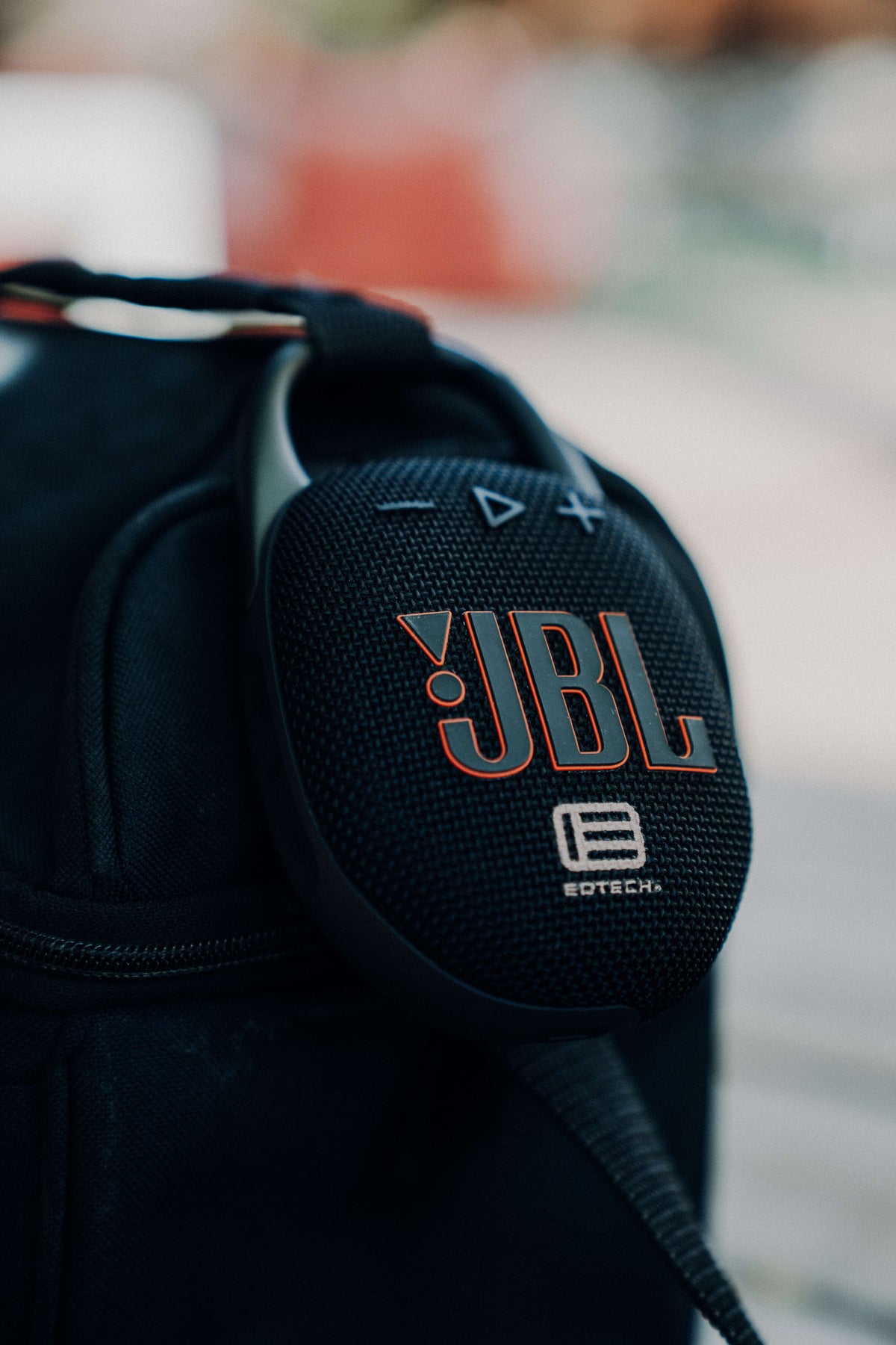 A close-up of a black JBL portable speaker bag with a round mesh-covered JBL logo patch, photographed outdoors against a blurred background.