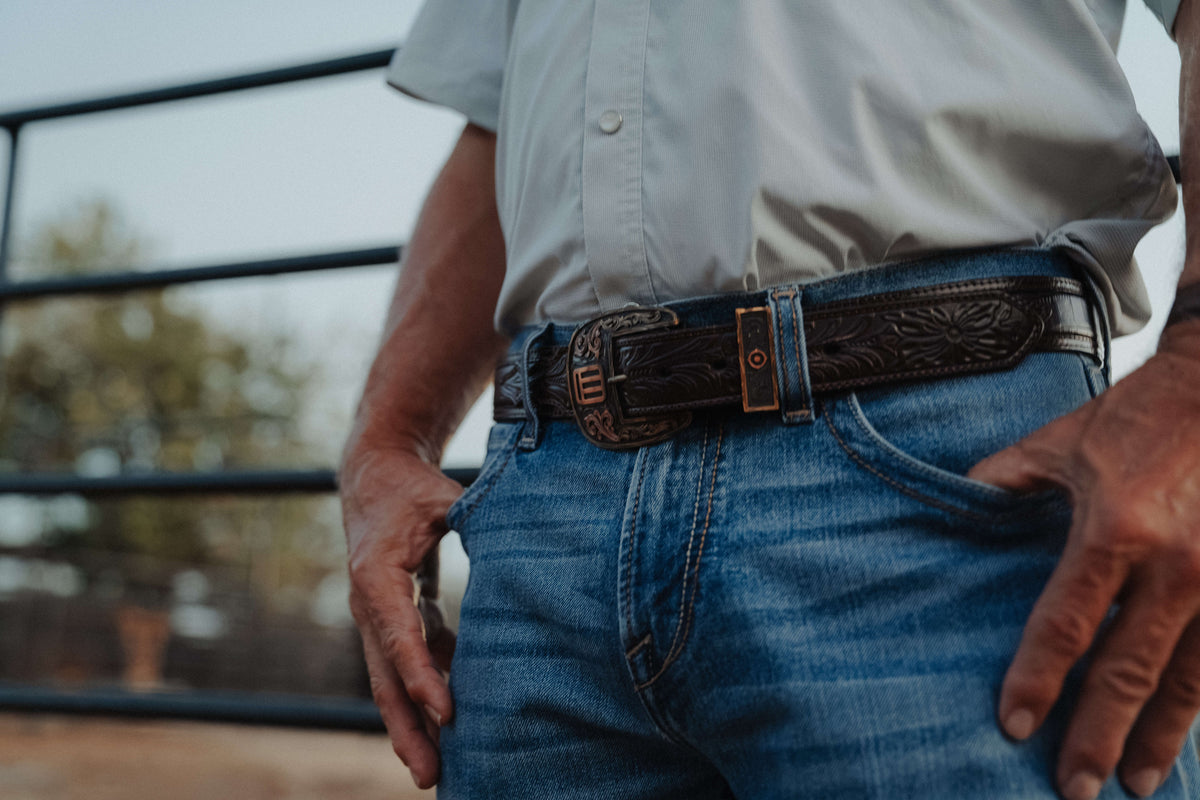 A close-up lifestyle shot of a man wearing a dark brown tooled leather belt with a bronze buckle, paired with blue jeans and a light gray shirt.